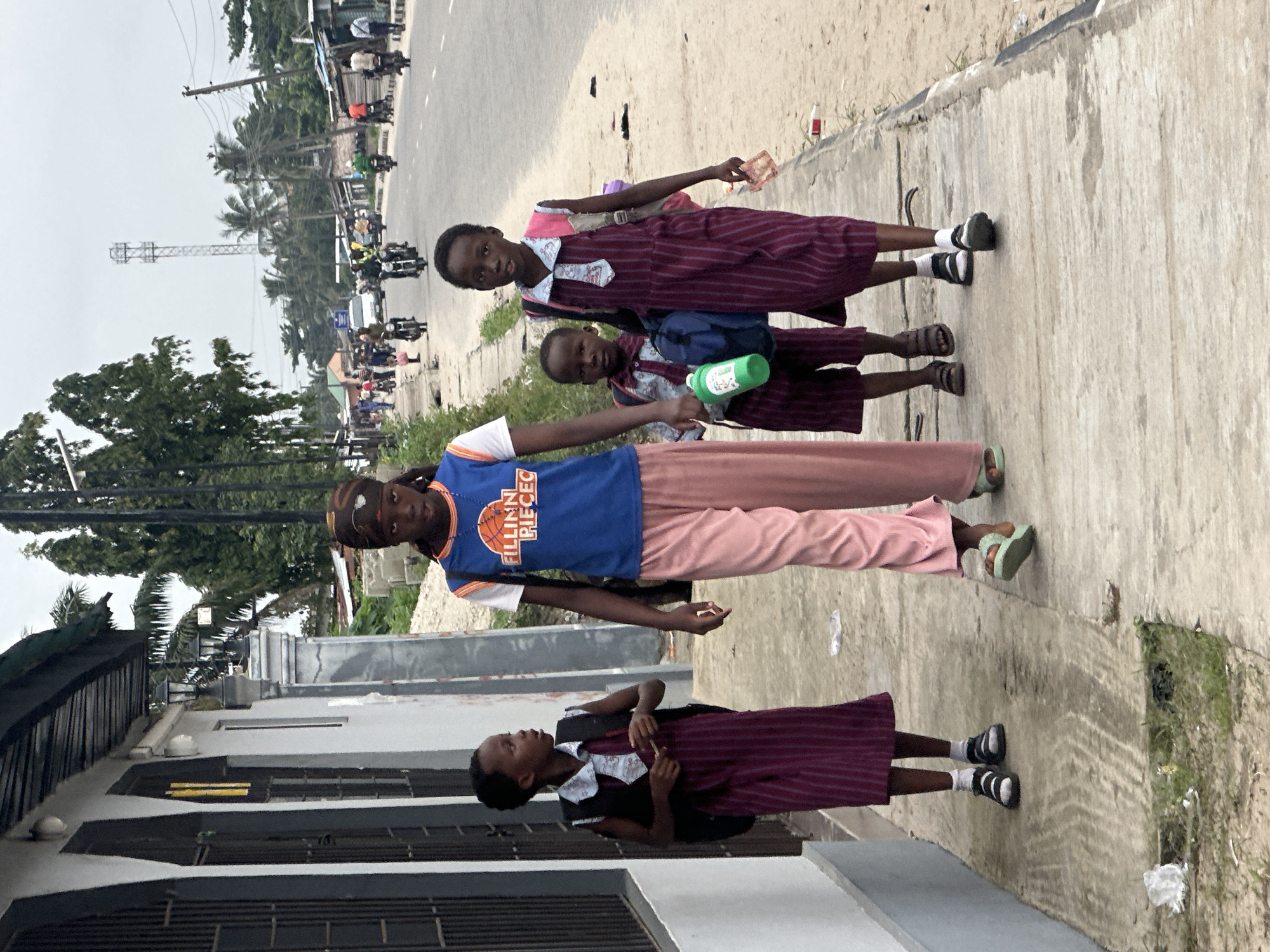 In Badagry, Nigeria, on a cloudy day, four young individuals, likely students, walk along a paved sidewalk. The sidewalk is bordered by a grey building on the left and a dirt road with a grassy verge on the right. The building features dark, arched doorways. On the road, several motorcycles and a few cars are visible, indicating a street with moderate traffic. Tall trees and palm trees line the background, with a telecommunications tower in the distance. The sky is overcast.
The individuals are walking from left to right. The leftmost person is a young girl wearing a dark purple dress with vertical stripes, a black vest, a patterned collar, and sandals. She carries a black backpack. Next to her is a slightly younger child, a boy, in a similar dark purple striped outfit with a patterned collar and carrying a blue backpack and a green water bottle. To their right, a girl in a blue t-shirt with an orange graphic and light pink pants walks with a young child. The child is wearing a dark purple striped dress and white socks with sandals. The oldest-looking individual, at the front and center, is wearing a blue t-shirt with the text "FILLINN PIECEG" and light pink pants, also walking towards the right. They all appear to be moving in the same direction, suggesting they are on their way to or from school. The overall scene depicts a common street view in a Nigerian urban environment, highlighting daily life and local infrastructure.