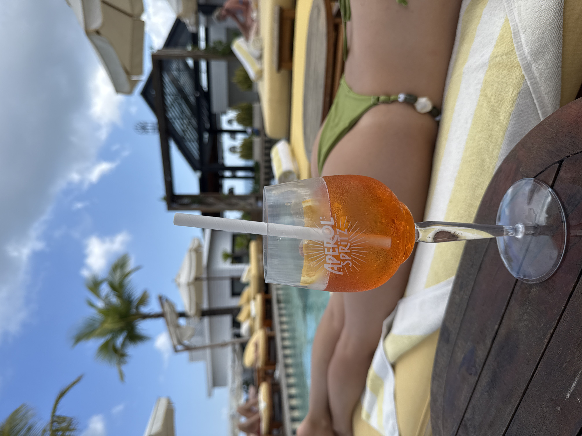 The image captures a vibrant scene by a pool in North Kuta, Indonesia. In the foreground, an Aperol Spritz cocktail sits on a dark wooden table. A white straw is partially submerged in the orange liquid. Behind the drink, a person lies on a sun lounger, their body partially visible, wearing a green bikini. A striped towel provides a soft backdrop. In the middle distance, the pool stretches horizontally. Around the pool, sun loungers with white umbrellas offer shade. Architectural features, including a pergola, are visible, with lush greenery adding to the tropical ambience. The sky is a clear blue, dotted with fluffy white clouds, and a palm tree stands tall on the left side of the frame.