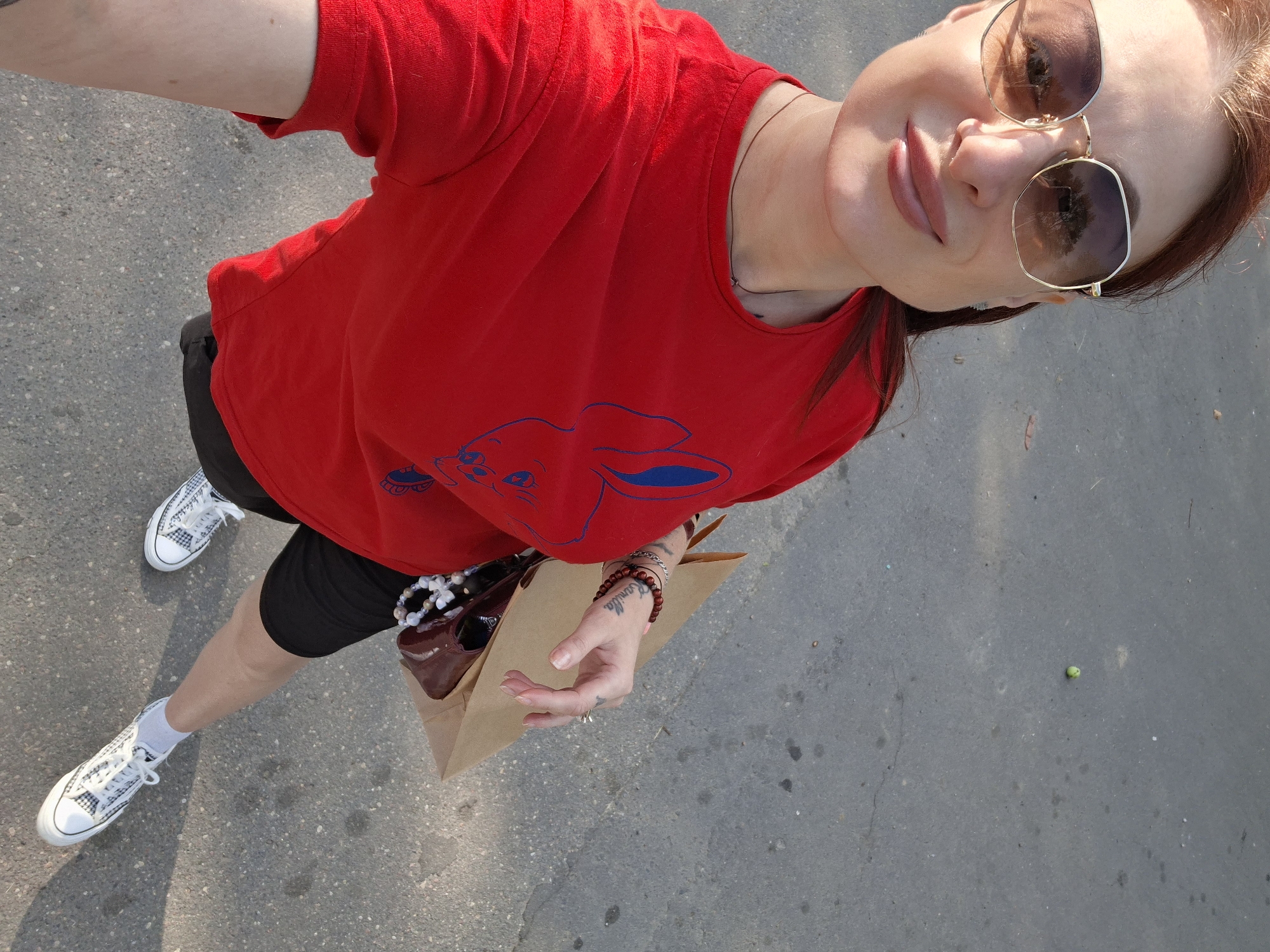 A woman with red hair and sunglasses is standing on an asphalt surface. She is wearing a red t-shirt with a blue bunny design, black shorts, white socks, and white patterned sneakers. She is holding a brown paper bag and a purse. Her left hand has tattoos and bracelets. The background is an outdoor environment with a grey asphalt ground. The location is identified as Забалоцкі сельскі Савет, Belarus.