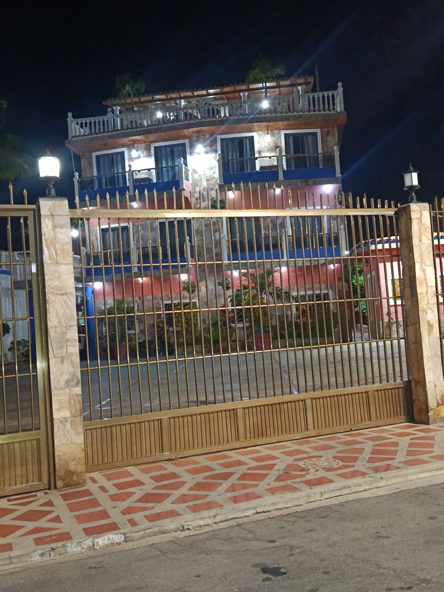 A three-story building is viewed through a gold-colored metal gate. The building has a stone facade, and each floor has a balcony with red and blue trim. The top floor has a white balustrade railing. The building is illuminated with several lights. Two lamp posts stand on either side of the gate. The sidewalk in front of the gate is paved with red and white tiles. The street is visible in the foreground. The time of day appears to be nighttime. The location is Juan Griego, Venezuela.