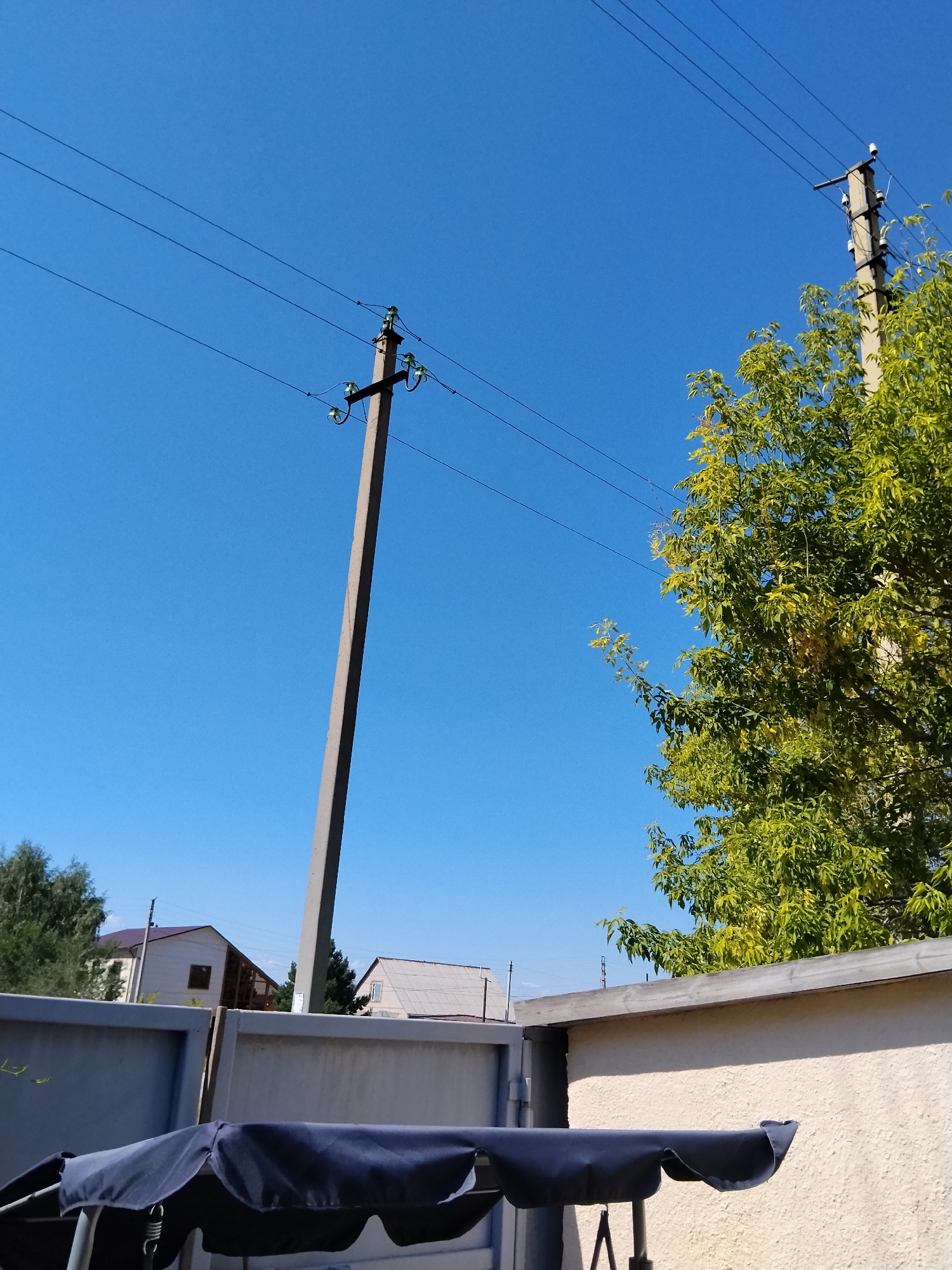 This is a low-angle, eye-level, medium shot of a residential area on a clear, sunny day. The sky is a vibrant, cloudless blue. In the foreground, a dark blue fabric canopy of a swing set is visible, partially obscuring a light-colored textured wall and a gray metal fence.
Dominating the mid-ground are two concrete utility poles. The pole on the left is tall and slender, angled slightly towards the right of the frame, with several sets of electrical insulators and wires. The pole on the right is shorter and sturdier, also supporting electrical wires. To the right of the right-hand pole, the dense, green foliage of a leafy tree extends into the frame.
In the background, beyond the fence and between the utility poles, are several residential buildings. The houses have pitched roofs, and one appears to be light beige with a dark-tiled roof and a visible window. Another house has a gray, corrugated roof. Trees with darker green foliage are scattered behind the houses.
The scene suggests a suburban or rural setting within the city of Kokshetau, Kazakhstan, likely a backyard or communal area with overhead power lines and residential structures. There are no people or animals visible in the image. The lighting indicates it is daytime, with the sun high in the sky.
