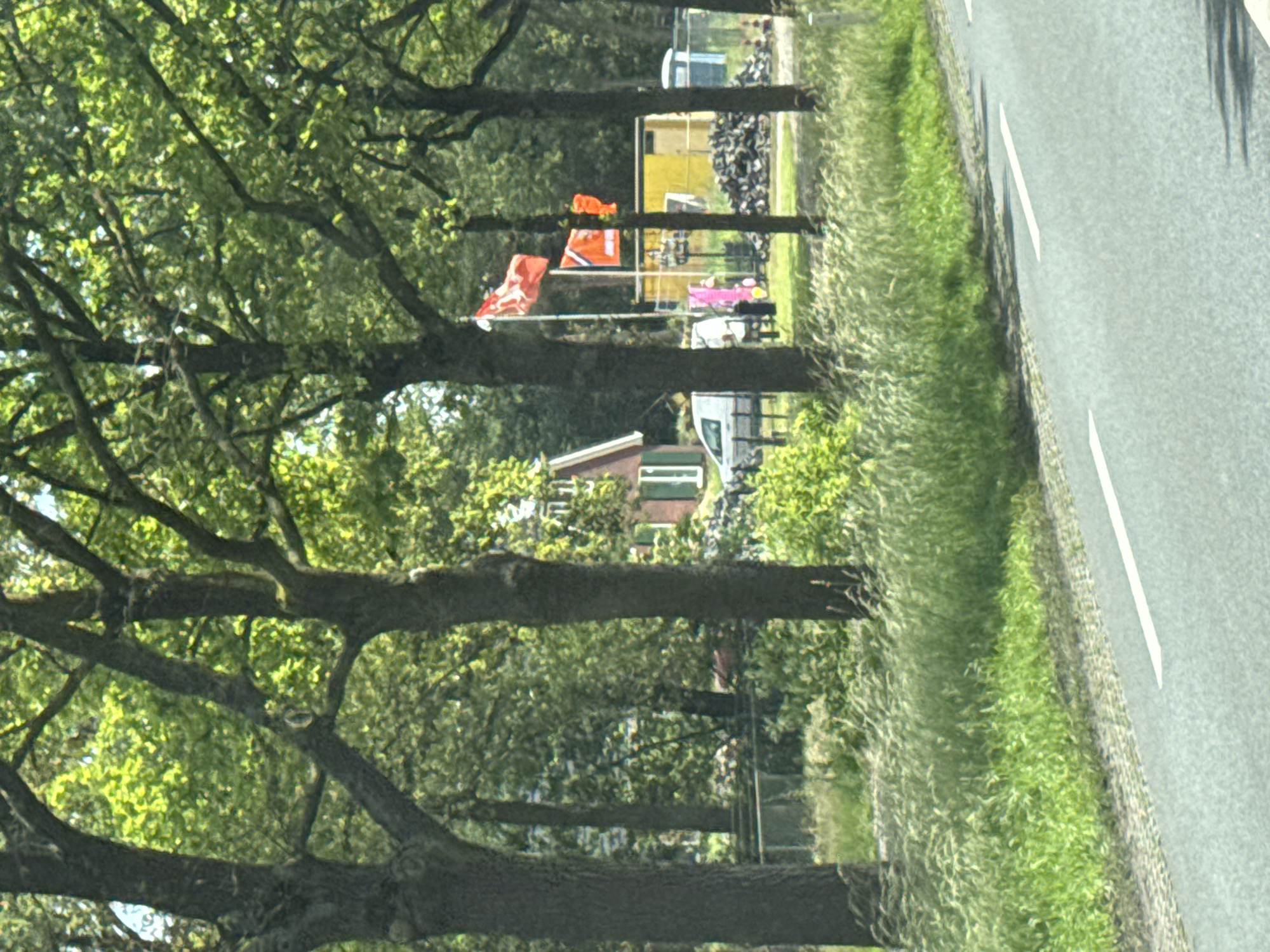 The image depicts a roadside scene in Wierden, Netherlands. In the background, behind a row of mature trees lining the road, a red-brick house with white trim is visible. Adjacent to the house are temporary structures, including a yellow portable building and a blue portable toilet. Two flags are displayed on poles in front of these structures: a red and white flag, and a solid orange flag. A white van is parked near the house. The foreground shows a paved road with white lane markings and a grassy verge with tall grass. The overall scene suggests a rural or semi-rural setting with some construction or event-related activity.