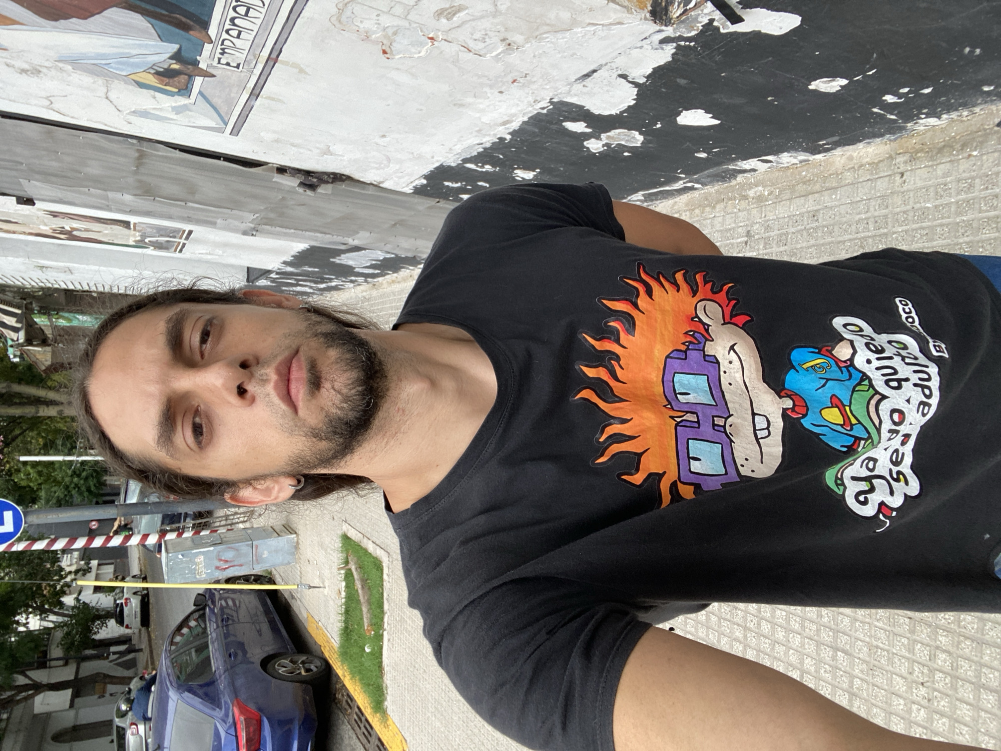A selfie of a man with long brown hair and a beard stands on a sidewalk, wearing a black t-shirt with Rugrats cartoon characters. A blue car is parked beside him. Behind him is a street with a striped railroad crossing arm. A mural is visible on the wall behind him, with the word "EMPANADAS" written on it. The photo appears to be taken in Buenos Aires, Argentina.