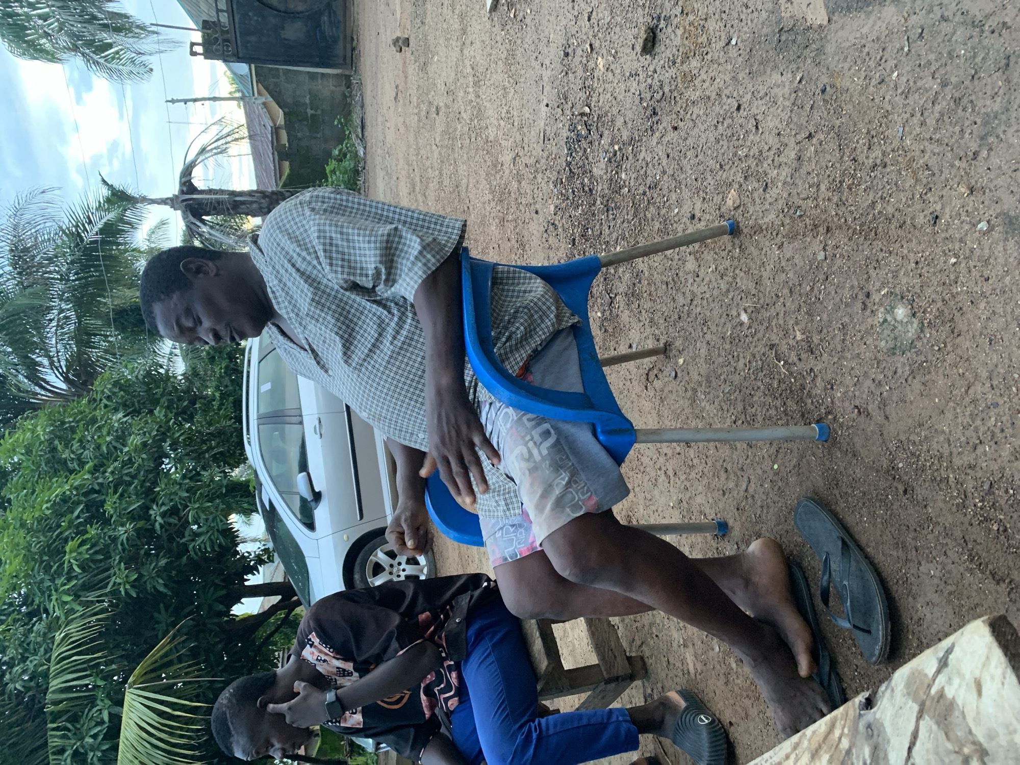 Two young men are sitting outdoors in a rural setting in Akwanga, Nigeria. The man in the foreground, wearing a checkered shirt and grey shorts, is seated on a blue plastic chair. He appears to be resting his head on his hand, with his eyes closed or downcast, suggesting a moment of relaxation or contemplation. Beside him, a second young man, dressed in a black shirt and blue pants, is seated on a wooden stool, looking down at his hands, which are near his face.
The ground is dusty and covered with small stones and debris. In the background, a silver car is parked, and lush green trees, including palm trees, surround the area. A simple building structure with a metal roof is visible behind the car. The lighting suggests it is daytime, possibly late afternoon, with diffuse light and a cloudy sky, indicating possibly overcast or humid weather. The overall scene conveys a sense of everyday life in a rural Nigerian community. No discernible text is visible in the image.