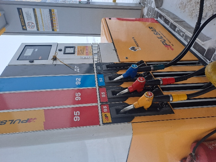 The image shows a gas station pump with four nozzles. From top to bottom, they are blue (92), red (95), and two yellow (95 and BP 95). Each nozzle is connected to a black hose. The pump has a digital display and price labels next to each nozzle. The structure is primarily gray, white, and yellow. Part of a fire extinguisher is visible at the bottom right of the image.