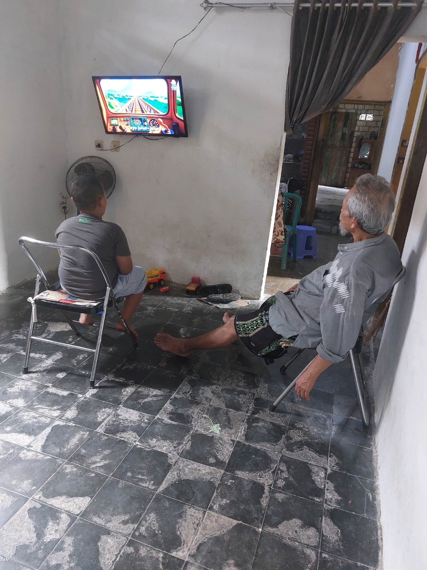 In a room in Sarangan, Indonesia, two individuals are seated, facing a television screen displaying a racing game. A younger person, viewed from behind, sits in a folding metal chair on the left, wearing a gray t-shirt and light-colored shorts. A small electric fan is positioned behind and to the left of this person. To the right, an older individual with gray hair and a beard is reclined in a similar folding chair, facing the television. This person is wearing a gray long-sleeved shirt and patterned shorts. Their left leg is extended forward. The room has white walls and a floor tiled with large, dark gray and lighter gray square tiles. A doorway covered by a dark gray curtain is visible on the right side of the frame, leading to another part of the building. Several toy vehicles, including a yellow dump truck and a red truck, are scattered on the floor near the younger person. The lighting suggests an indoor setting, possibly during the daytime.