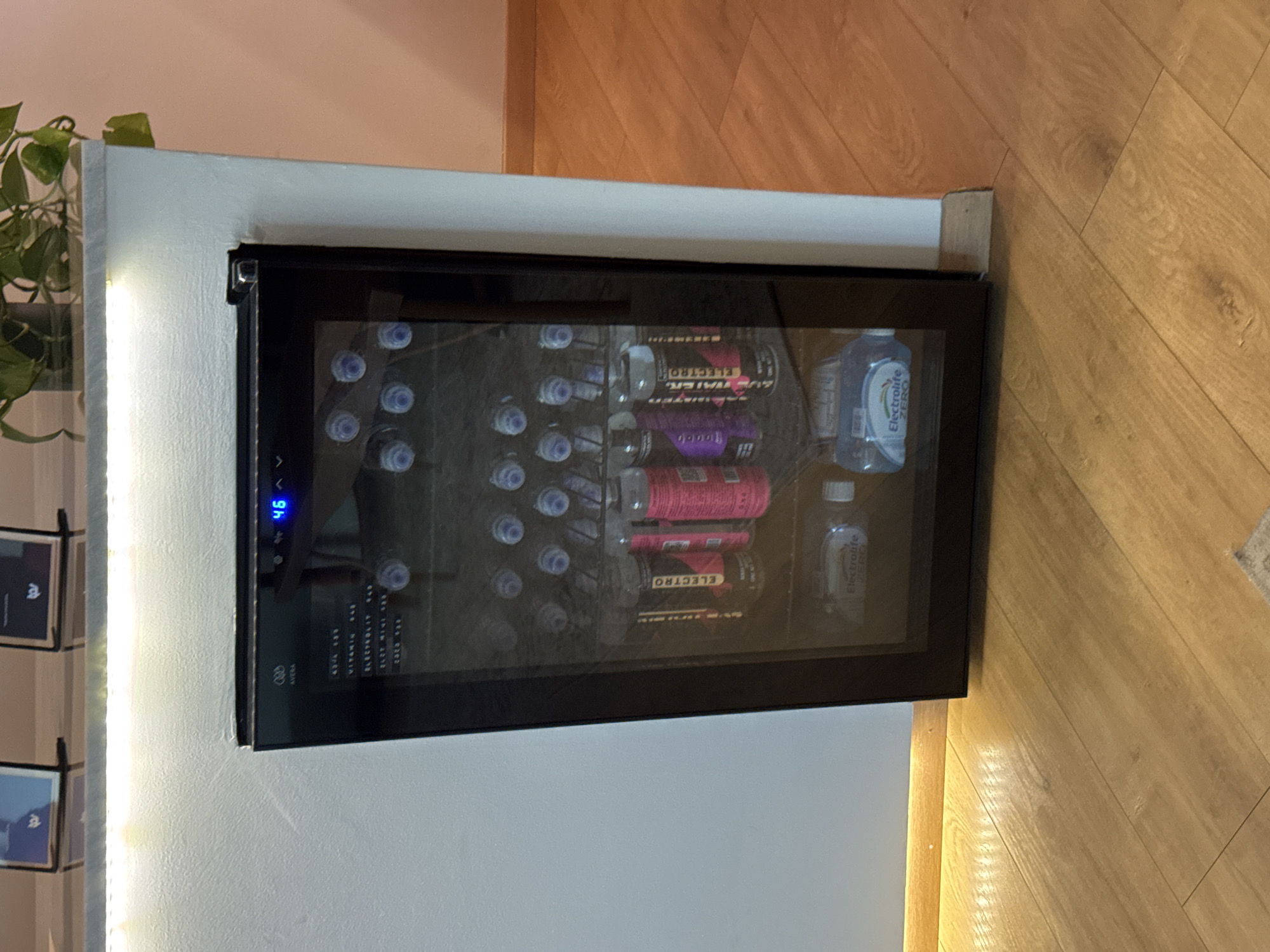 A compact, built-in beverage refrigerator is positioned against a light-colored wall and sits atop a white baseboard. The refrigerator, framed in black, is filled with bottled water and Electrolit drinks of various flavors. The digital display on the refrigerator shows a temperature of 46 degrees. To the left, there's a shelf with framed artwork and a green plant. To the right, the refrigerator meets a light brown wooden floor. The image was taken in Puebla, Mexico.