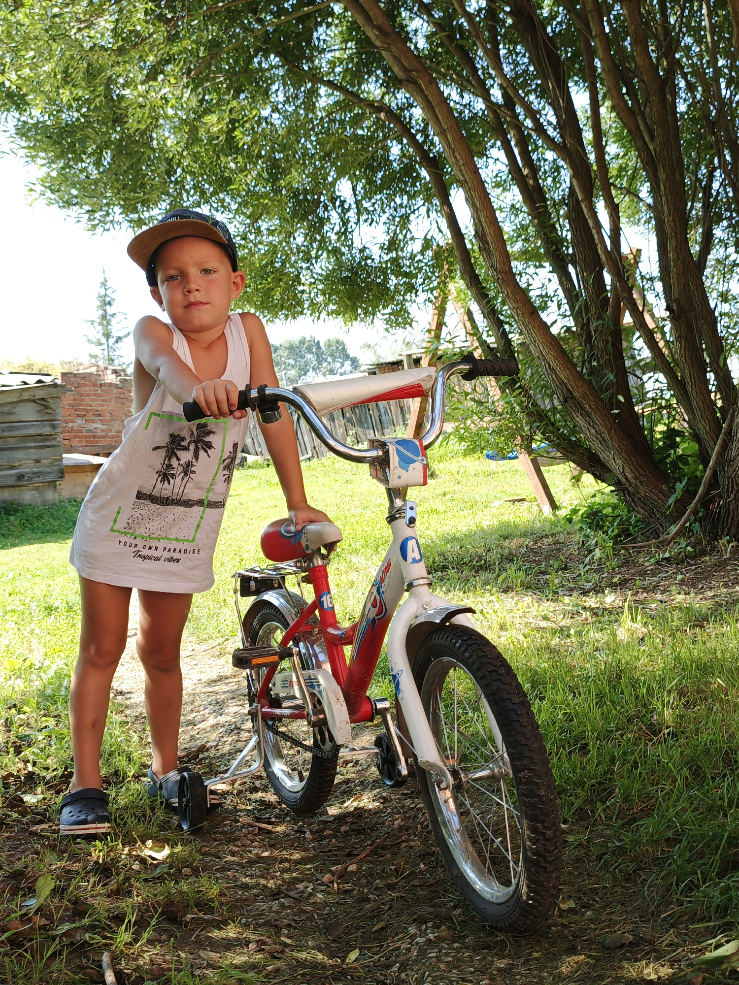 In the village of Medvedyovka, Russia, a young boy stands with his bicycle. The boy, wearing a white tank top with a tropical print and a baseball cap, leans on the handlebars of a red and white children's bicycle. The bicycle has training wheels attached. The scene is set outdoors, with lush green grass and the dappled shade of trees. In the background, a brick structure and some wooden fencing are visible, suggesting a residential or rural setting. The lighting indicates it is daytime, likely a sunny day with some overcast conditions filtering the light.