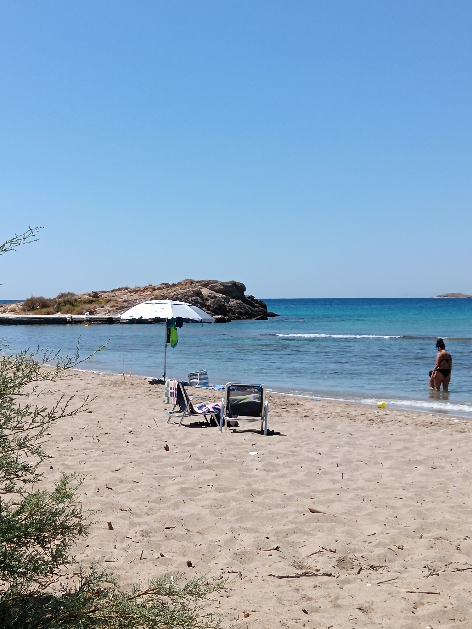 This is a full-frame, eye-level, outdoor shot of a sandy beach in Agia Marina, Greece, on a clear, sunny day. The shot is taken from the shore looking out towards the sea. The water is a vibrant blue, with gentle waves lapping against the shore. In the distance, there is a rocky island with some sparse vegetation.
In the foreground, the sandy beach is mostly empty, with a few scattered pieces of driftwood. There are two beach chairs and a large white umbrella set up near the water's edge. A towel is draped over one of the chairs. Further back, a woman in a black bikini is standing in the shallow water, facing away from the camera. Another person, partially submerged in the water, is visible behind her. A small yellow object, possibly a toy, is in the sand near the water.
To the left of the frame, a bushy green plant with thin branches partially obscures the view. The sky is a clear, bright blue with no clouds. The overall scene depicts a tranquil day at the beach.