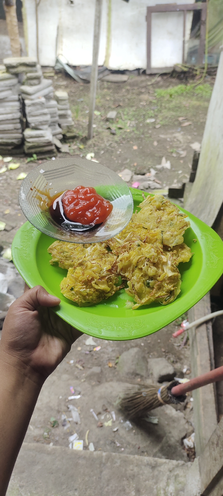 A person's hand holds a bright green plastic plate filled with crispy, golden-brown fritters, likely bakwan, a popular Indonesian vegetable fritter. A small, clear glass dish sits on the plate, containing a dollop of vibrant red chili sauce and a swirl of dark brown sauce, possibly soy sauce or sweet soy sauce. The fritters appear to be made with shredded vegetables, possibly cabbage or carrots, and are lightly battered and fried to a crisp. The background is blurred, suggesting an outdoor setting, possibly a rural area or backyard, with a rough concrete ground, some stacked bricks or stones, and the base of a building or wall. A broom with a red handle is also visible in the lower right portion of the image, partially sweeping the ground. The overall scene depicts a casual presentation of Indonesian street food.