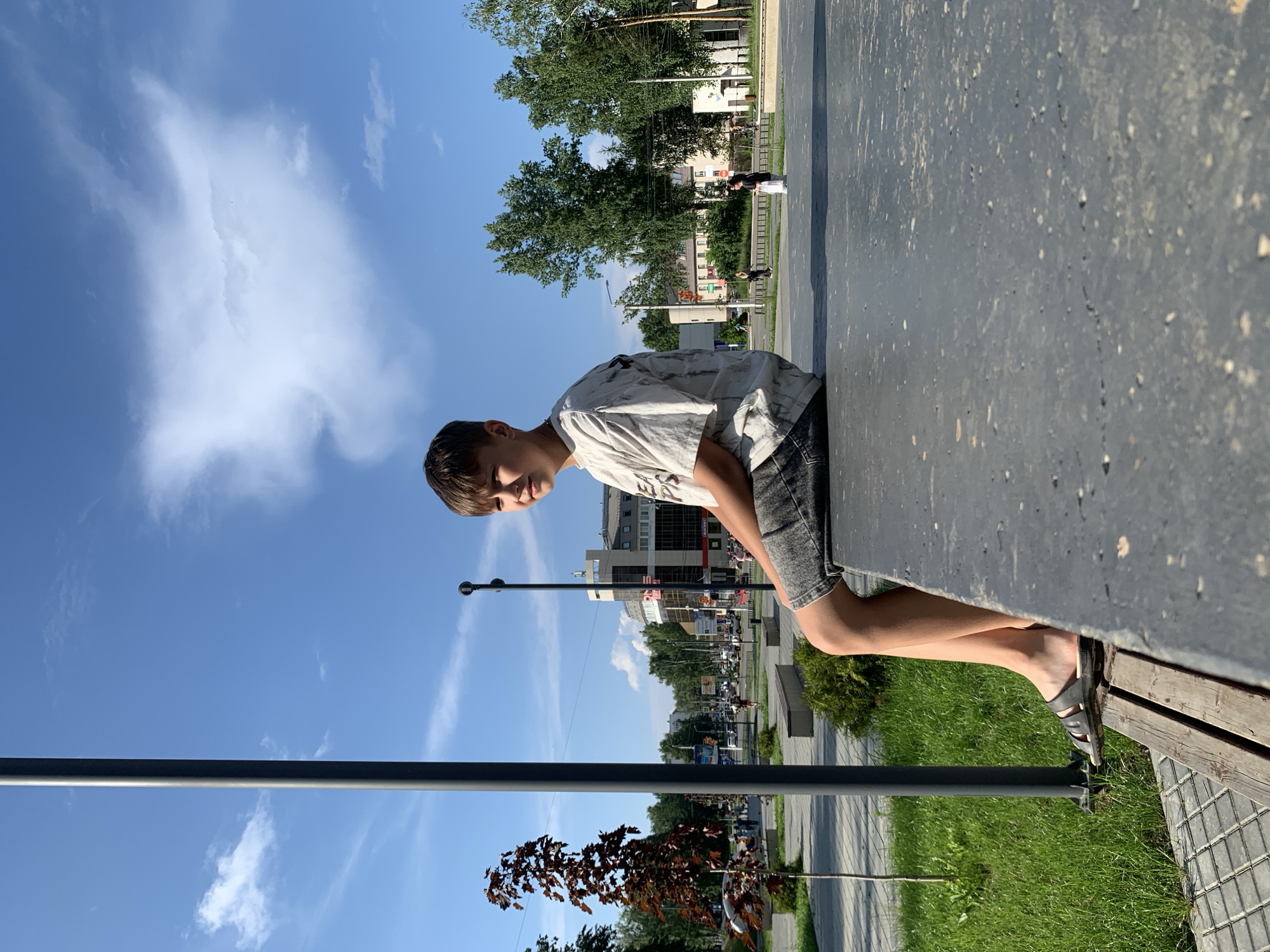 A young Caucasian boy with short, brown hair sits on a raised, gray concrete ledge in a public outdoor space, facing to the right. He is wearing a light-colored t-shirt with a dark, abstract pattern and dark gray shorts. His legs are bare, and he is wearing dark sandals on his feet. The boy is looking slightly upwards and to the right with a neutral expression.
The background suggests an urban environment in Chaikovsky, Russia. To the left, a tall, dark gray pole stands in front of a small, reddish-leafed tree and a paved walkway. Beyond that, a street with cars and buildings is visible. The buildings are modern, with glass facades and balconies, suggesting commercial or residential use. Green trees line the street and the background.
To the right of the boy, the concrete ledge continues, and in the middle ground, there is a paved area with people walking and riding bicycles. Further in the distance, more trees and buildings are visible. The sky is bright blue with scattered white clouds, indicating a sunny day. The lighting suggests it is late afternoon. The overall scene is a typical urban park or plaza setting.
