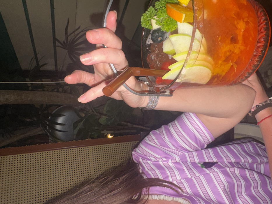 A medium close-up photograph taken at night in Ban Ko Li Pe, Thailand, shows an individual holding a large, clear glass beverage container. The person's right arm and torso are visible; they wear a light purple and white striped short-sleeved shirt with a dark fruit emblem on the left chest, a white beaded or ruffled neckline, a silver-colored mesh bracelet on the right wrist, and multiple dark beaded and red string bracelets on the lower right forearm.
The beverage container, resembling a fishbowl with a silver-colored wire handle, is filled with an orange-colored liquid and ice cubes. It is garnished with several slices of green apple, an orange segment, a sprig of green parsley, small dark berries (likely blueberries), and pieces of red fruit (likely strawberries). A brown plastic straw is inserted into the drink, held by the individual's right hand.
The background is dark, indicating a nighttime setting. Visible elements include dark tropical foliage, a light-colored woven rattan screen or partition on the left, and a dark, ribbed, inverted bowl-shaped object (likely a hanging light fixture or speaker) in the upper-center. A thin white cable runs vertically in the background. The scene depicts a leisure activity, likely at a restaurant or bar.