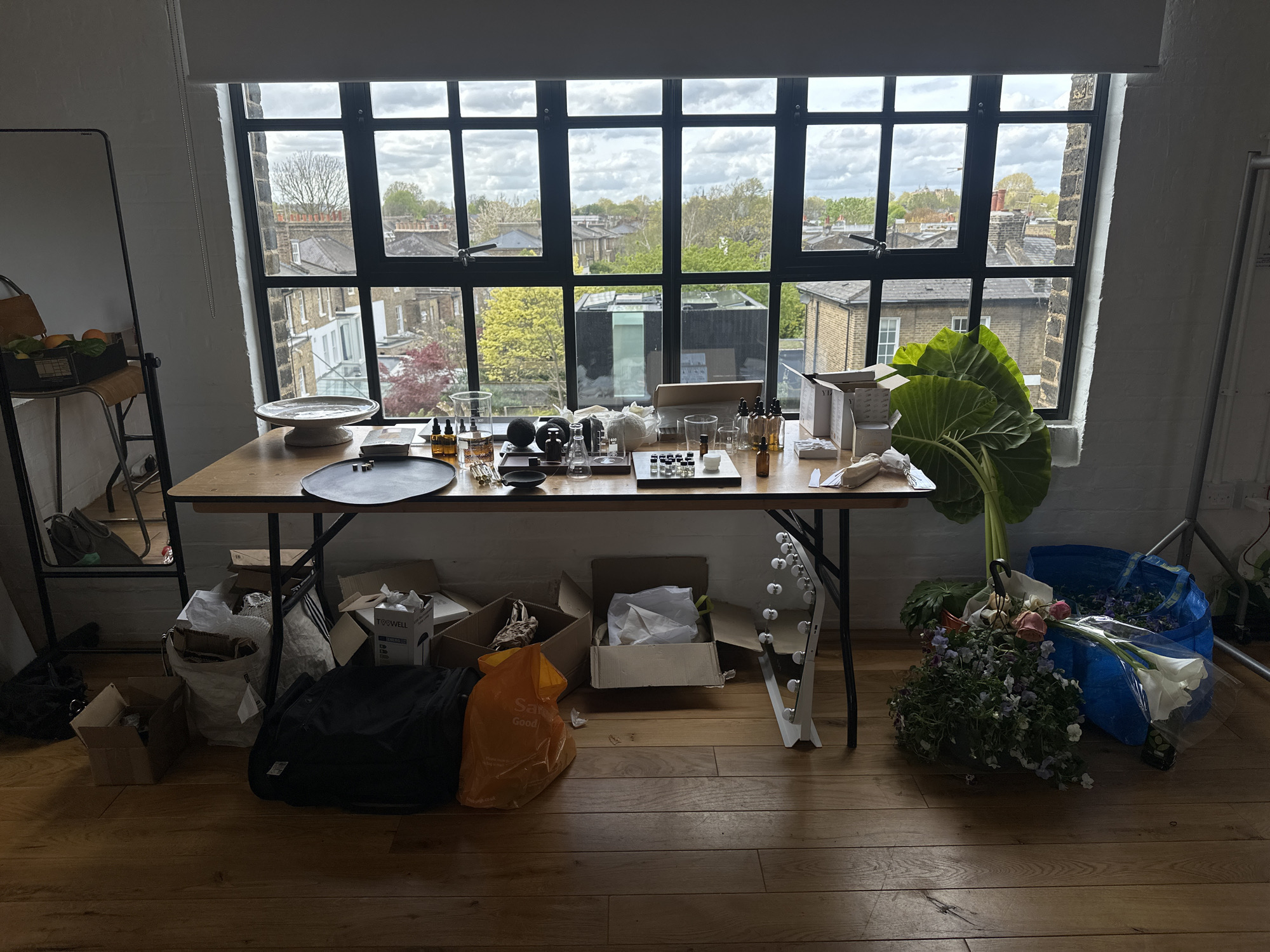 The image presents an indoor studio or workshop space, characterized by a large, multi-paned dark-framed window that overlooks an urban landscape. Through the window, various brick buildings, rooftops, and trees are visible under an overcast, cloudy sky, consistent with a daytime setting in a city like London, United Kingdom. The room features a light wooden floor and white brick walls, suggesting a converted industrial or loft space.
A long wooden table spans the width of the window, serving as a workbench. It is laden with numerous objects, including various glass bottles with droppers, beakers, small vials, and white ceramic containers, indicating activities related to product creation, possibly perfumery or skincare. Other items on the table include a black circular tray, small black bowls, a white ceramic cake stand, and several open cardboard boxes, some marked "Y.B." A prominent feature on the right side of the table is a cluster of large green leaves, resembling elephant ear plants.
Underneath the table, several cardboard boxes, some containing packaging materials (one box says "TOSWELL"), a black duffel bag, and an orange plastic bag with "Sainsbury's Good" printed on it are arranged. A white storage rack with circular holders also sits below the table. To the far left, a tall rectangular mirror leans against the wall, beside a chair holding a basket of fruit, likely oranges or lemons.
On the wooden floor to the right of the table, a substantial pile of fresh flowers, including roses, pansies/violets, and calla lilies, lies wrapped in paper and plastic. Two large blue IKEA Frakta bags are nearby, one appearing to hold more foliage or flowers.
The scene suggests an active creative workspace, possibly involving floristry and product formulation, conveying a sense of preparation or ongoing work. No people are visible. The overall ambiance is industrious under natural, diffused light.