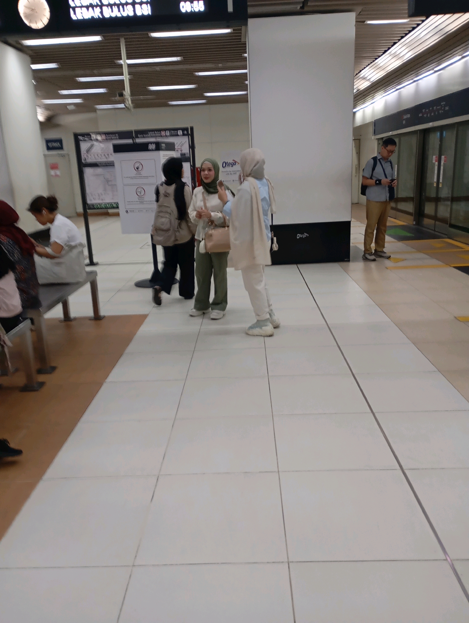 In Central Jakarta, Indonesia, a person stands near a subway platform with a man in the background looking at his phone and a subway train. In the foreground, two women are talking. The platform is clean with tiled flooring and a digital sign above.