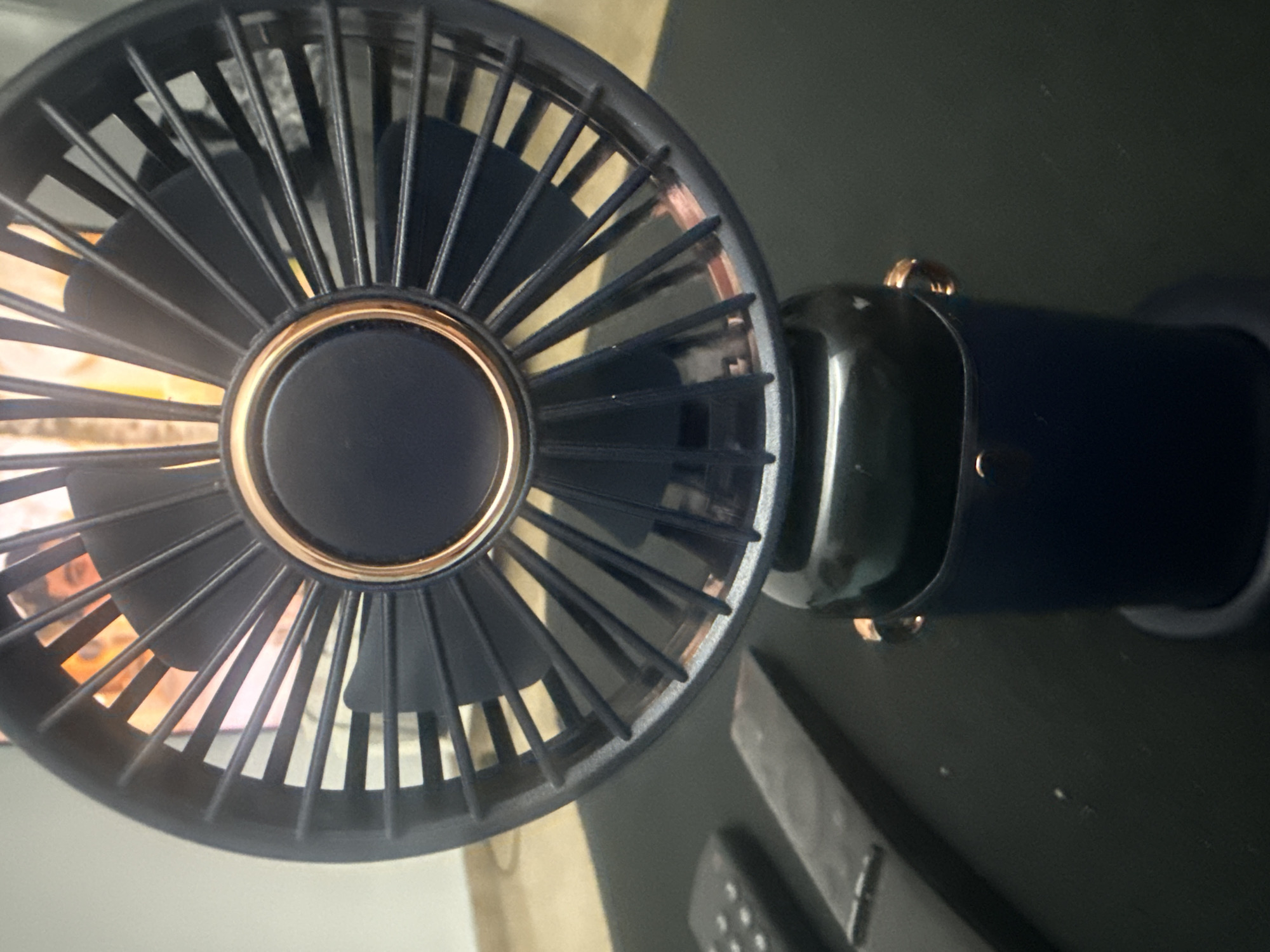 This is a close-up, eye-level, indoor shot of a dark blue portable fan with a metallic rose gold accent ring around the center. The fan is positioned on a dark, textured surface, likely a desk or table. To the left of the fan, out of focus, are two dark rectangular objects, one appearing to be a remote control with several buttons. Behind the fan, a person's face and shoulders are visible, slightly out of focus, with warm lighting illuminating them, suggesting a screen or light source. The background is a plain, light-colored wall. The scene is devoid of any specific landmarks or indicators that would allow for pinpointing a location within Valencia, Spain.
