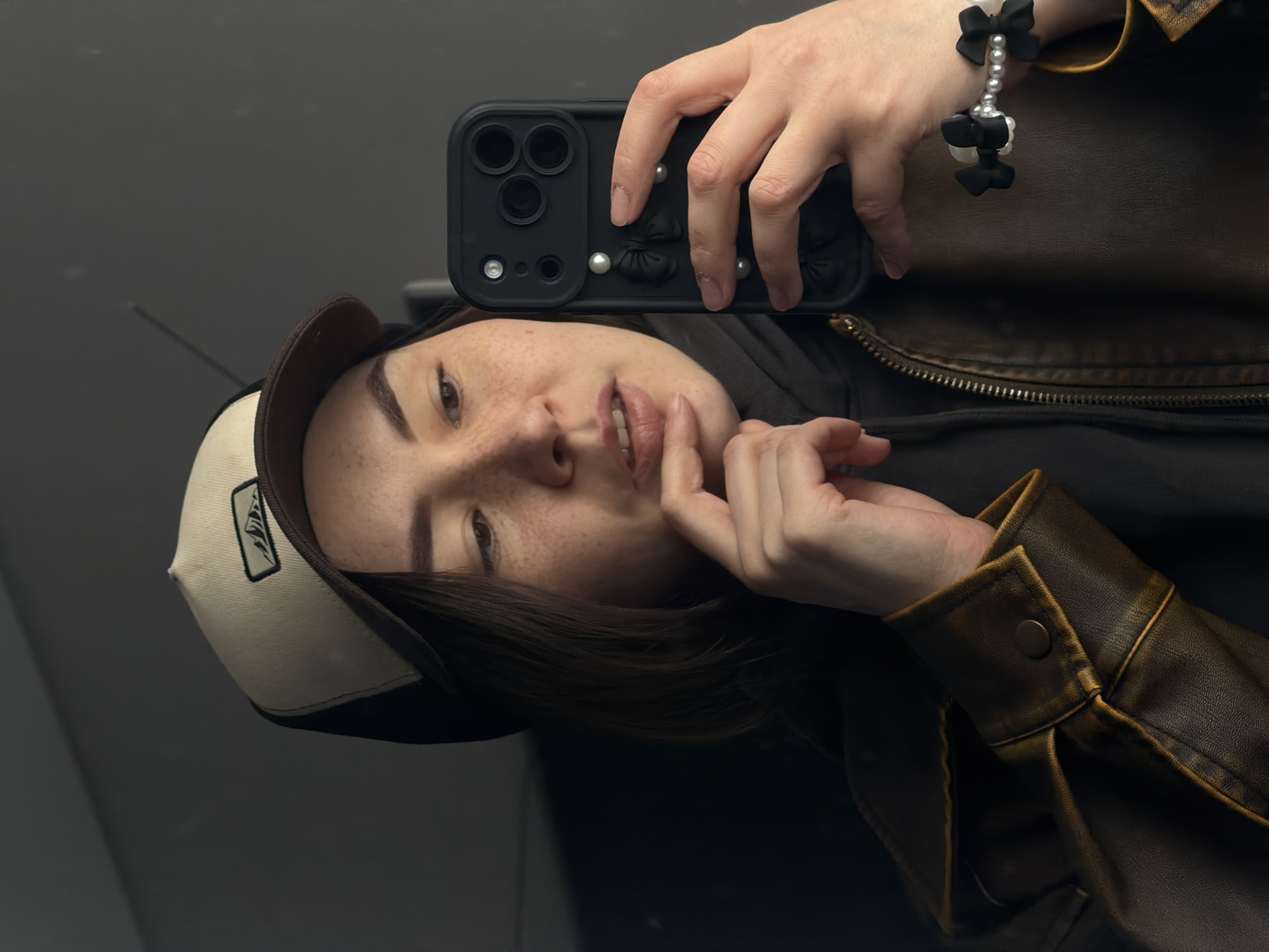 A young woman takes a selfie using a black smartphone with a decorative case featuring bows and pearls. She is wearing a black and tan baseball cap with a logo, a black hooded sweatshirt, and a brown leather jacket. A bracelet with black bows and pearls adorns her wrist. The photo appears to be taken indoors, possibly in Chaikovsky, Russia, although no definitive landmarks confirm this location. The background is a dark, reflective surface.