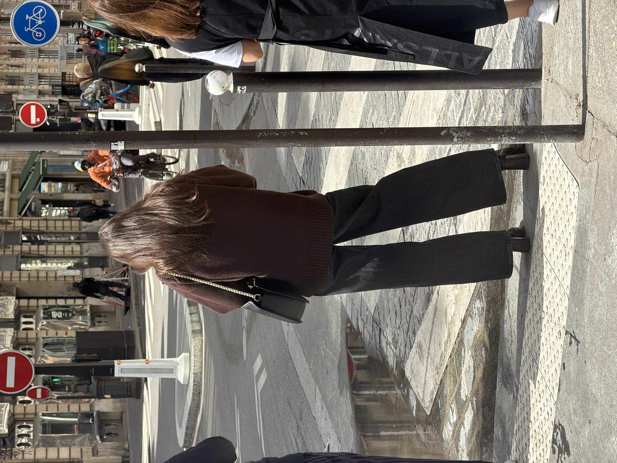 The image captures a vibrant street scene on a sunny day in a city, likely Paris. The perspective is from slightly behind two individuals, looking down a bustling street with typical Parisian architecture.
In the foreground on the right, a person with long, dark brown hair is visible from the back, wearing a brown knitted sweater and dark wide-leg trousers. They carry a small, dark shoulder bag with a gold-colored chain strap. To their left, another person with brown hair wears a black jacket over a white top and dark pants, paired with white sneakers. Faint text, possibly "SALLE" or "BALLE," is discernible on the back of their jacket.
A series of dark metal poles or barriers runs across the mid-ground, separating the foreground from the street. Beyond these, the street features patterned paving stones on the sidewalks and white line markings on the asphalt road, including a crosswalk. Several individuals are present: a cyclist wearing an orange jacket and a helmet is visible, along with other pedestrians.
Notable street signs include two red circular "No Entry" signs on the left side of the street and a blue circular sign with a white bicycle symbol, indicating a bicycle route or area. In the background, multi-story buildings with classic stone facades, numerous windows, and balconies line the street, characteristic of Parisian urban landscapes. The bright sunlight casts clear shadows, suggesting it is daytime with good weather. The overall activity depicts a typical day in a lively city environment.