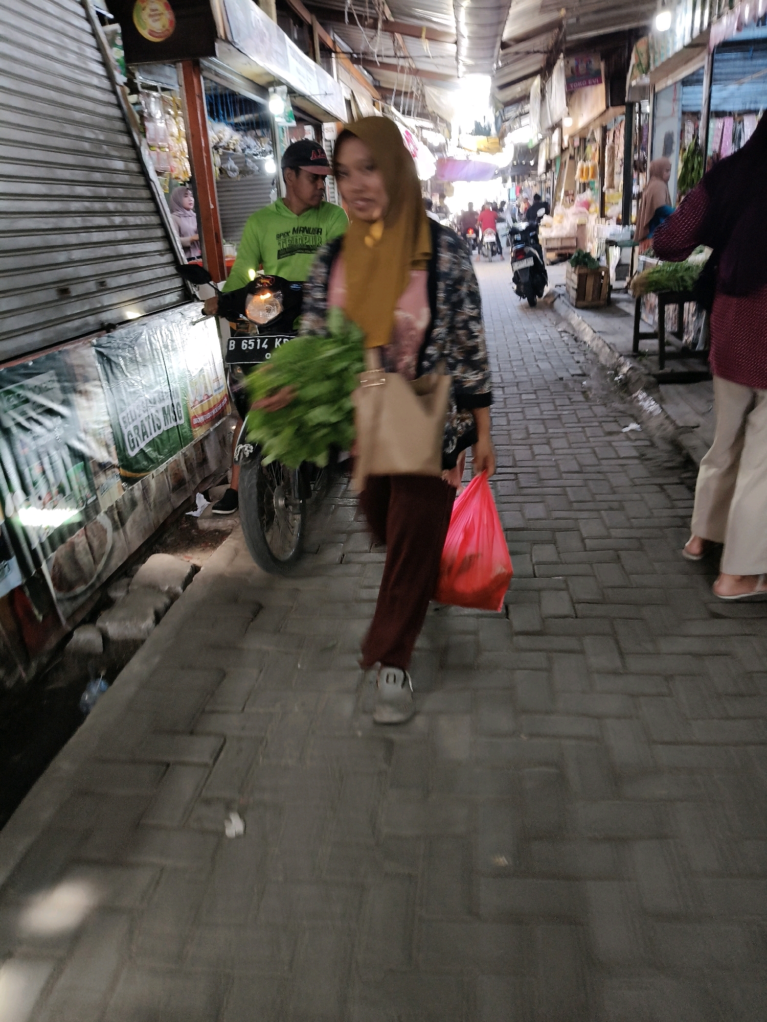 This is a vertical, eye-level, full shot of a busy street market in Setiamekar, Indonesia. The foreground is dominated by a woman walking towards the camera, carrying a red plastic bag in her right hand and a beige tote bag on her left shoulder. She is wearing a floral-patterned jacket over a pink shirt and brown pants. Her face is out of focus, but she is wearing a mustard-colored hijab. To her left, a man on a motorcycle, wearing a green hoodie and a black baseball cap, is looking away from the camera. The motorcycle has a license plate that reads "B 6514 KG". Behind the man, a store with rolled-down shutters is visible, and a woman in a pink hijab is peeking out from it. The market street is paved with grey brick pavers. On the right side of the image, another woman is walking away from the camera, wearing a maroon top and cream-colored pants. She has a bouquet of green vegetables in her left hand. The street is lined with market stalls and buildings, some of which have awnings or roofs made of corrugated metal and fabric. In the background, more people and motorcycles are visible, and the market extends into the distance.