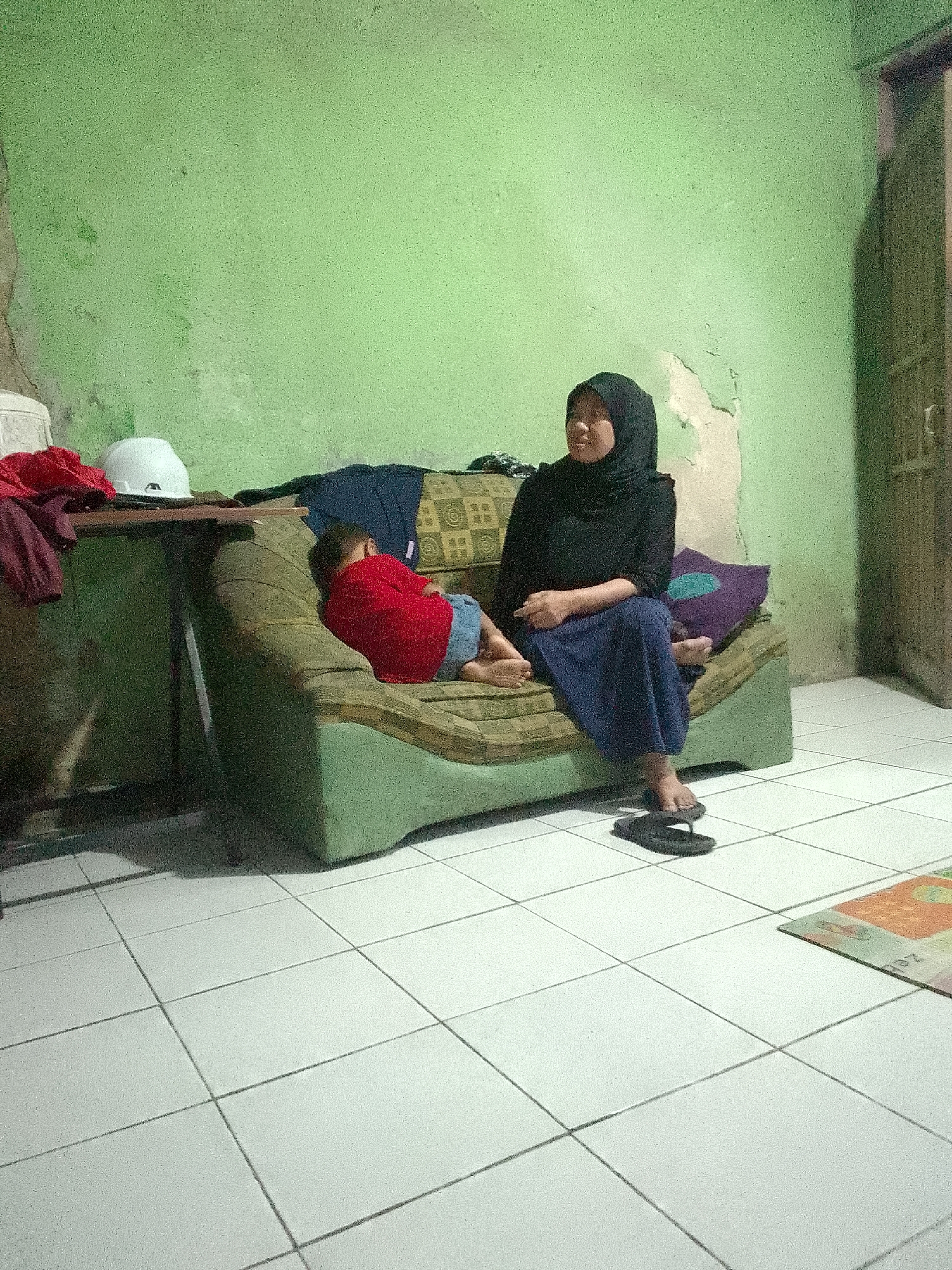 A woman wearing a black hijab and blue pants sits on a green sofa in a room with a green, textured wall. A child in a red shirt and blue shorts lies on the same sofa, facing away from the camera. Next to the sofa, to the left, is a table with a white helmet and some red clothing. To the right of the woman, a purple pillow with a teal pattern rests on the sofa. The floor is tiled with white squares. A pair of black flip-flops lies on the floor in front of the sofa. The room appears to be a residential interior, possibly in Gunungmasigit, Indonesia.
