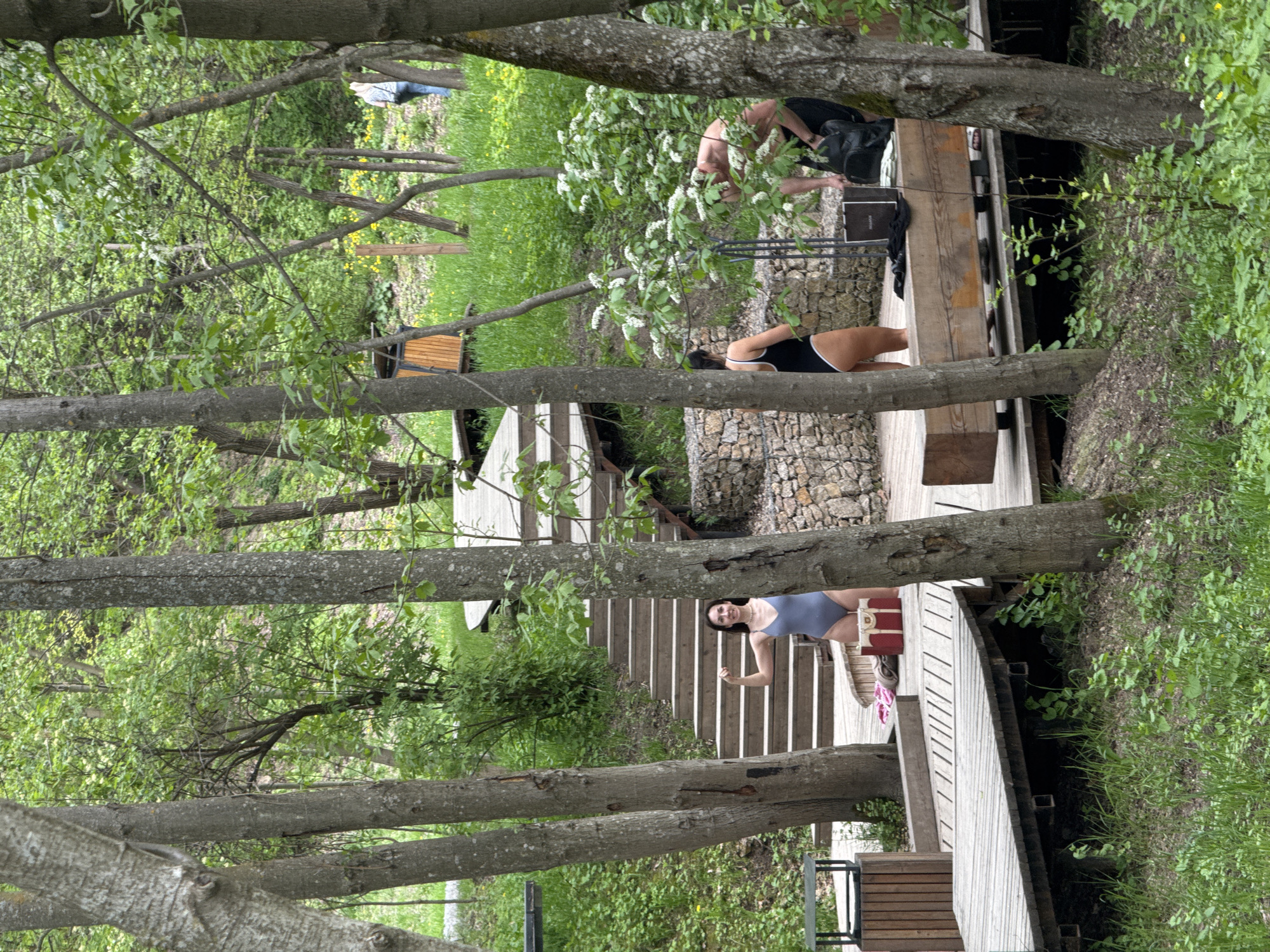 The image depicts a partially obscured view of a park-like setting in Sdt Shcherbakovka, Russia. Several individuals are present at different points along a wooden walkway and adjacent stone-lined area. In the foreground, a woman in a blue swimsuit stands on a wooden platform with stairs behind her. She is near a suitcase and pair of pink flip-flops. Further up the walkway, another woman in a black and white swimsuit is visible. Beyond her, a man is bent over near some bushes. The walkway appears to cross a stream or water feature. The scene is framed by numerous tree trunks and foliage, which partially obscure the view.
