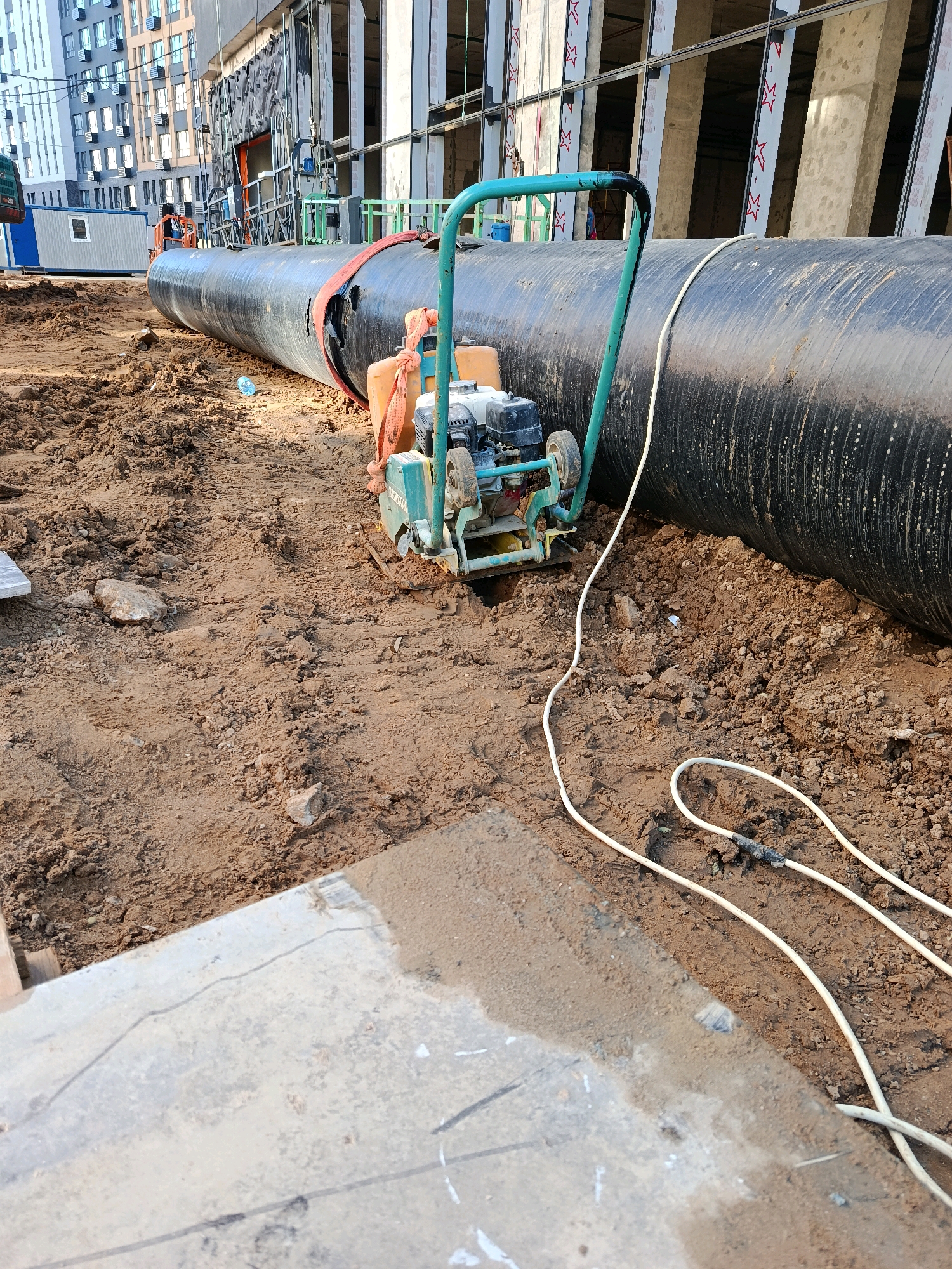 In Vnukovo, Russia, a construction site displays a large black pipe, possibly for underground utilities, resting on a bed of dark brown soil. The pipe is secured with an orange strap. In the foreground, a vibratory plate compactor, painted in turquoise and featuring an orange engine cover and a metal handle, sits on the dirt. A white electrical cord snakes across the ground from the compactor, disappearing into the soil. Beyond the pipe, the skeletal framework of a modern building with concrete pillars and large windows under construction is visible, alongside two modular buildings. The ground is uneven, with loose soil, rocks, and a patch of concrete in the lower left corner of the frame.