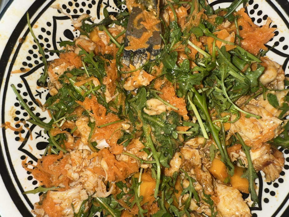 A colorful salad in a patterned bowl features shredded carrots, chunks of mango, and what appears to be shredded chicken. Fresh arugula adds a peppery note. The salad seems tossed with a dressing that coats the ingredients, giving them a glossy sheen. A utensil, possibly a fork, is visible amidst the salad, indicating it's ready to be eaten. The bowl's design adds a decorative element to the presentation.
