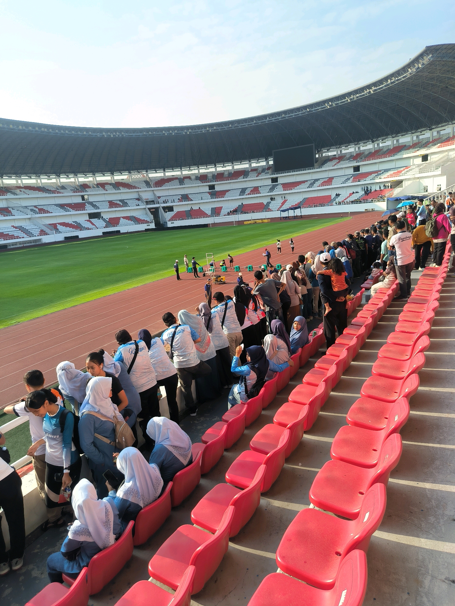 The image depicts an outdoor stadium in Semarang, Indonesia, likely during a sporting event or gathering. The stadium features tiered seating in red and white, with a large, green field visible in the background. A running track encircles the field. A considerable number of people, many wearing light-colored t-shirts and some wearing hijabs, are seen standing and sitting in the spectator seats. Some are looking towards the field, while others are engaged in conversation. The lighting suggests it is daytime, with sunlight casting shadows across the seating area. The architecture of the stadium, including its arched roof structure and the design of the seating, is characteristic of modern sports venues.