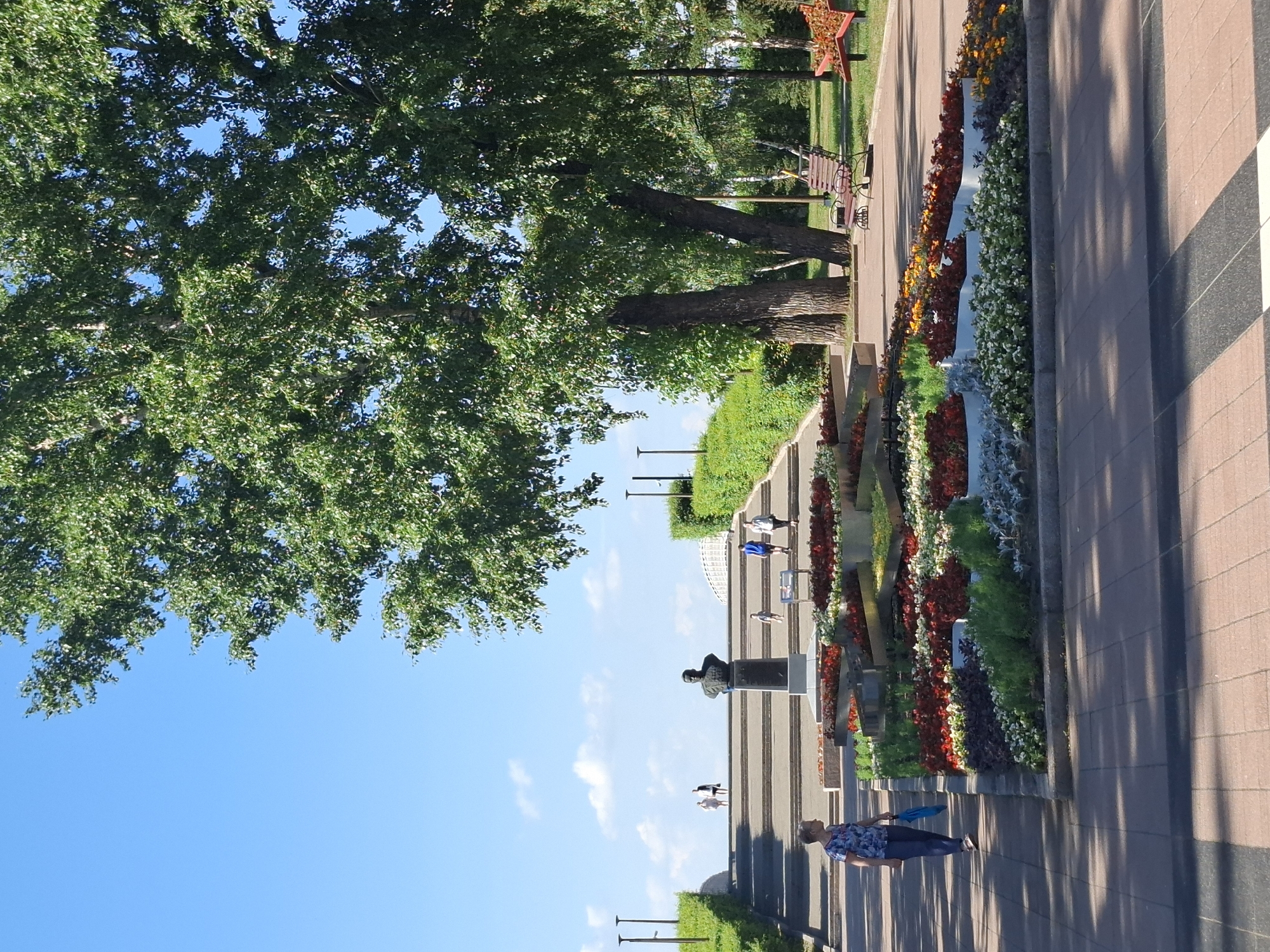 The image shows a sunny day in a park in Irkutsk, Russia. The scene is outdoors and features a walkway lined with colorful flowerbeds and several trees. A statue of a man in a suit is prominently displayed on a pedestal along the walkway. Several people are strolling along the path, some are standing, and some are walking in groups. One person is holding a blue object that might be a fan or an umbrella. The time of day appears to be afternoon, given the long shadows cast by the trees and the angle of the sun. The sky is clear and blue, indicating pleasant weather. The overall atmosphere of the scene is relaxed and leisurely, suggesting a typical day in a public park. There is no visible text in the image.