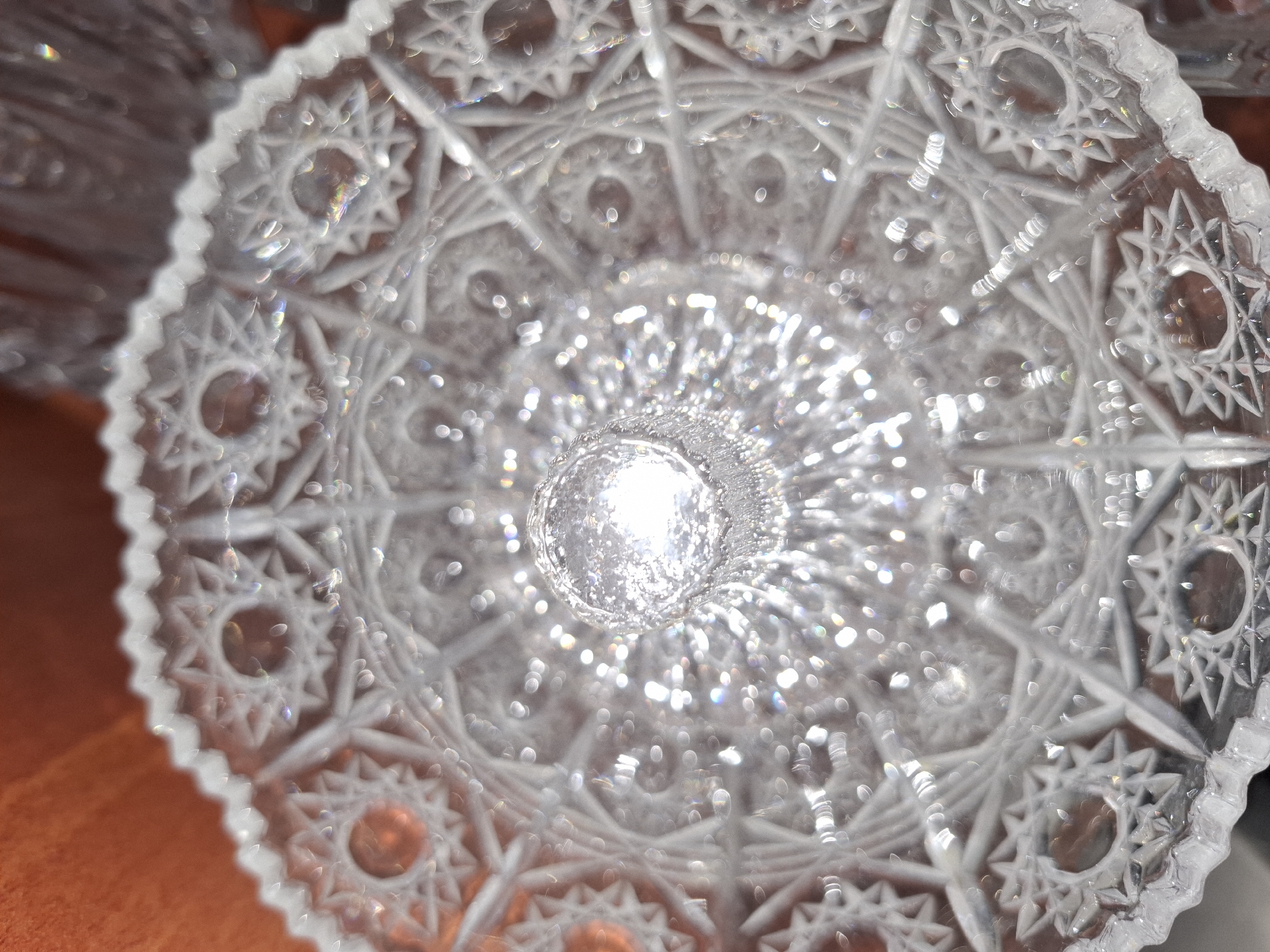 The image is a close-up, top-down view of a cut crystal glass object, possibly a bowl or a lid, resting on an orange surface. The crystal is intricately patterned with geometric shapes, stars, and radiating lines, which refract light, creating numerous bright highlights and some rainbow-like color flares. A central raised element, also made of cut crystal, is visible in the middle of the object, with a bright light source reflecting intensely from its surface. The overall impression is one of sparkle and opulence, with the glass appearing highly polished and detailed. The orange surface beneath the crystal has a soft, textured appearance, possibly fabric or a blurred background. There are no people, animals, or any identifiable landmarks or features of the city of Almaty, Kazakhstan visible in this image. The context of Almaty is entirely absent from the visual information presented.