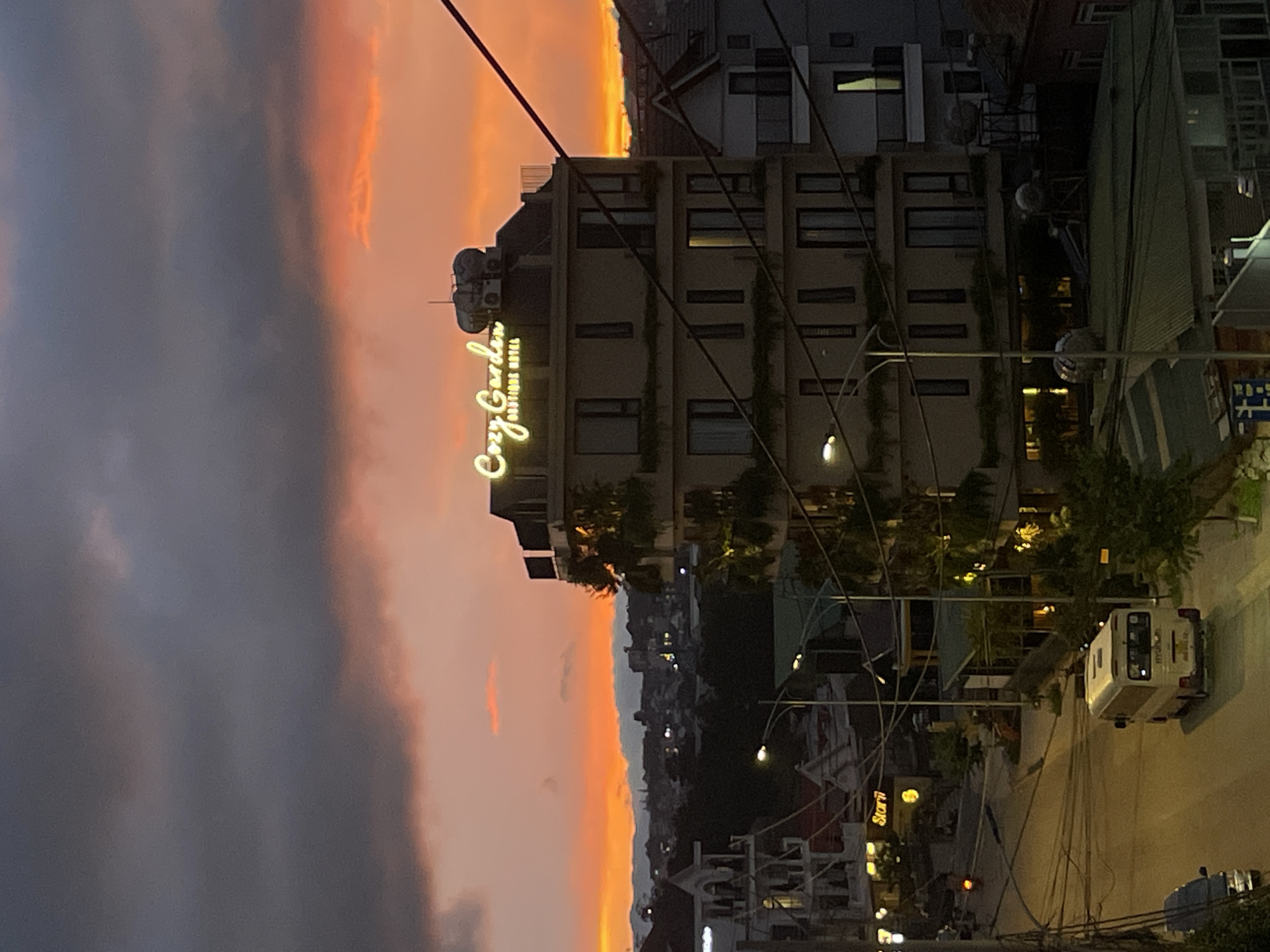 An image captures a street scene in Lâm Viên - Đà Lạt, Vietnam during sunset. The sky is ablaze with vibrant orange and red hues, contrasted by dark, ominous clouds. In the mid-ground, a multi-story building with a lit "Cozy Garden Boutique Hotel" sign stands prominently. Greenery is visible cascading from balconies on the building. In the foreground, a white minivan is parked on the street, facing away from the viewer. A dark-colored car is parked to the left of the minivan. Streetlights illuminate the road, which curves to the left. The background shows a silhouette of a city with scattered lights and distant mountains. Electrical wires crisscross the frame, obscuring parts of the sky and buildings. The overall atmosphere is one of urban twilight with a dramatic natural backdrop.