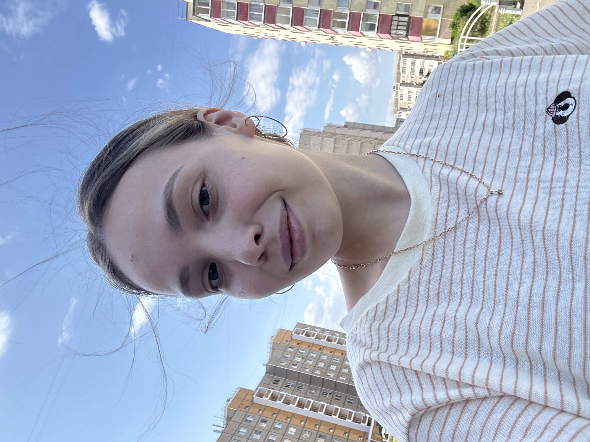 A young woman with light brown hair, wearing a white and peach striped shirt, is taking a selfie in what appears to be Chaikovsky, Russia. She has a gold hoop earring and a delicate gold necklace. The backdrop features residential buildings of various heights and architectural styles, under a partly cloudy blue sky.