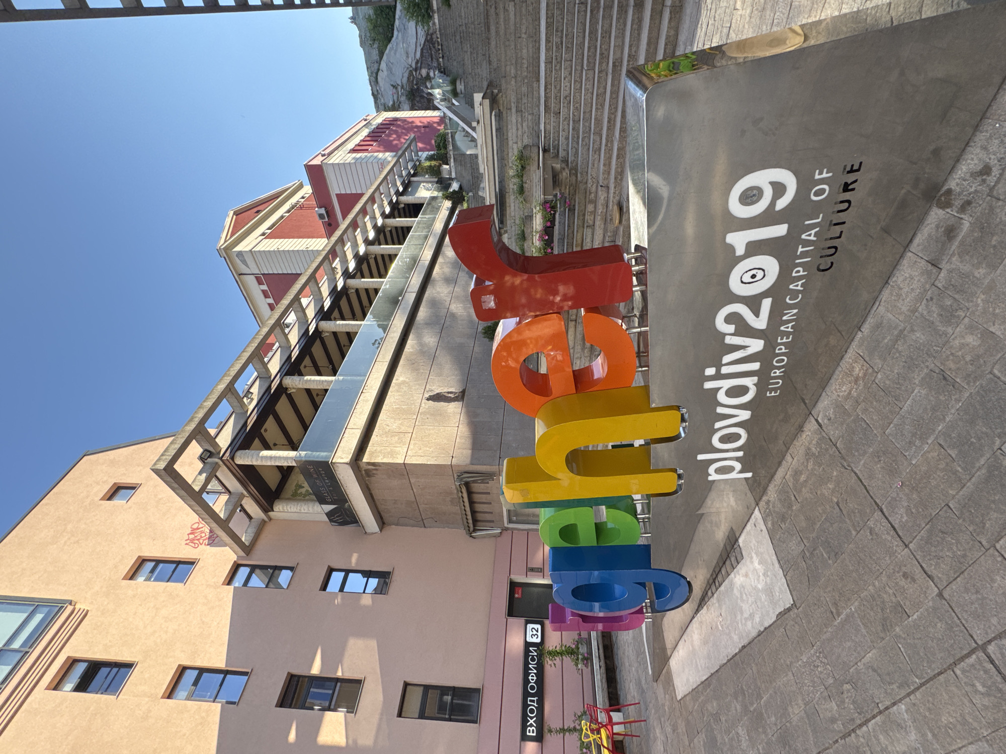 The image shows an outdoor scene in Plovdiv, Bulgaria, featuring a large, rainbow-colored sign that spells out "Plovdiv2019 European Capital of Culture." The sign is situated on a paved area, likely a plaza. Behind the sign is a multi-story building with a balcony. Adjacent to the building is a staircase leading uphill towards a building with a red roof. In the background is a clear blue sky. The lighting suggests it's daytime, possibly late morning or early afternoon. A doorway is visible with "Вход Офиси 32" written on a sign, which translates to "Entrance Offices 32."