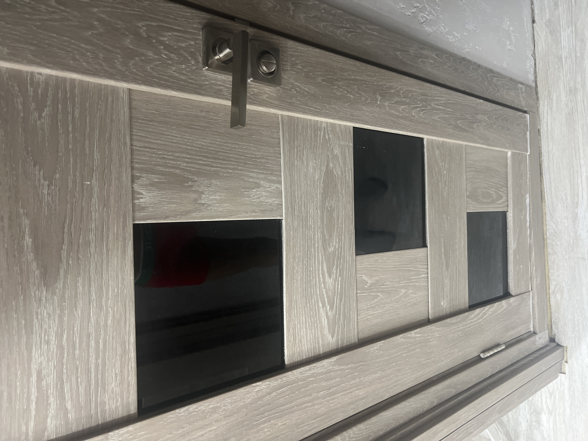 A close-up, low-angle shot focuses on a light brown wooden door with a contemporary design. The door is divided into several panels, some of which are fitted with rectangular black glass inserts. A modern, brushed-metal door handle is visible on the right side of the door. The wood grain is pronounced, with a whitewashed finish that highlights the natural texture. The surrounding environment suggests an indoor setting, with a glimpse of a wall on the right and a light-colored floor at the bottom, both appearing to have a similar wood-grain texture or finish. A person's legs, wearing red and green patterned shorts, are partially visible through the top black glass insert. The image lacks any clear indicators of its specific location within Veteran-2, Russia, beyond it being an interior scene with a door.