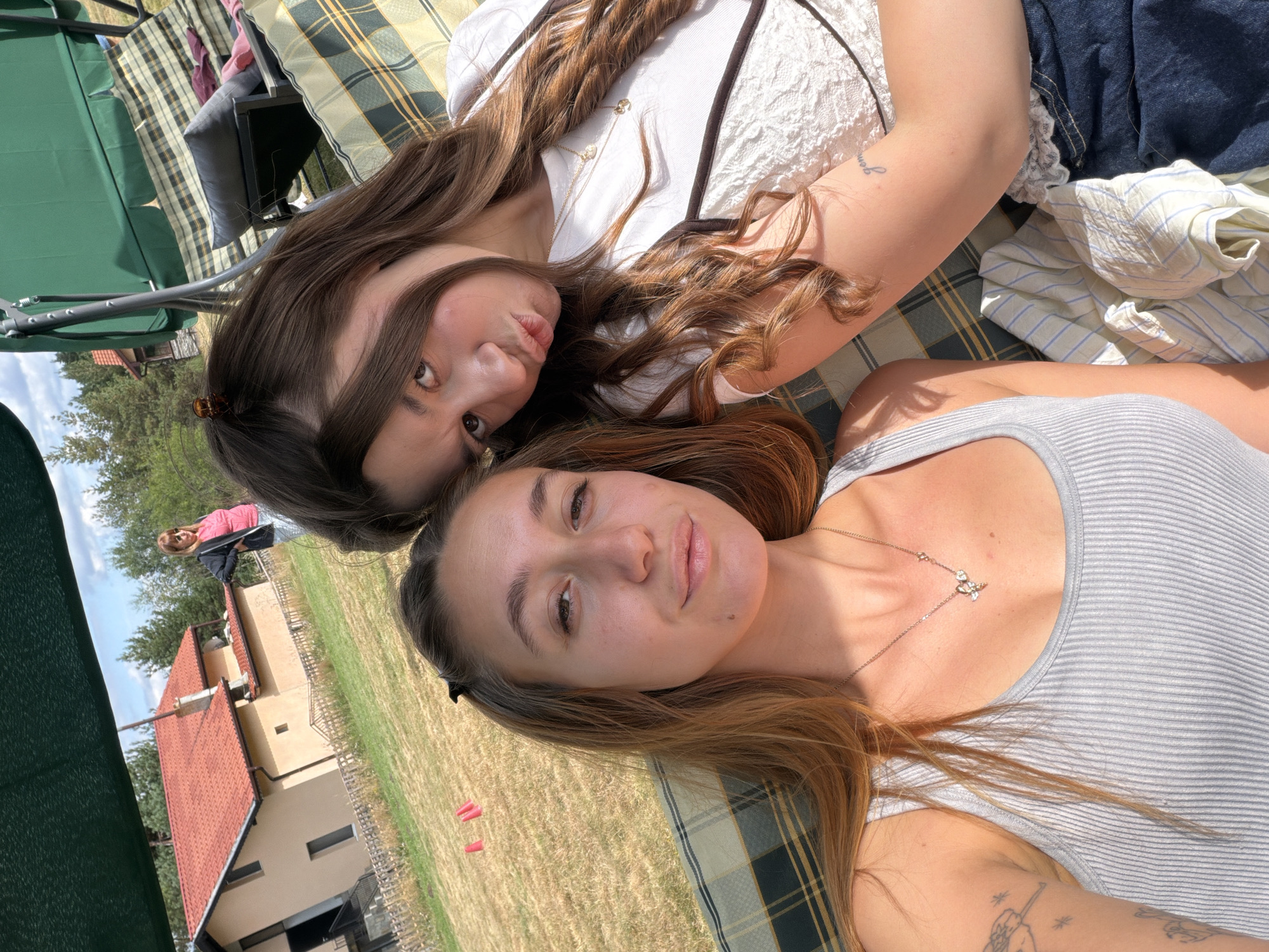 Two young women are captured in a close-up selfie, seated on a patterned cushion of an outdoor swing. The woman on the left, with auburn hair, wears a grey ribbed tank top and a delicate butterfly necklace. The woman on the right, with long brown hair, is wearing a white lace top and has her hair styled with a clip. She is puckering her lips towards the camera. In the background, a tan house with a red tile roof is visible, situated in a grassy area surrounded by trees, suggesting a rural setting. A woman in a pink shirt and dark jacket stands near the house. The overall scene appears to be a sunny day in the city of Galabovo, Bulgaria.