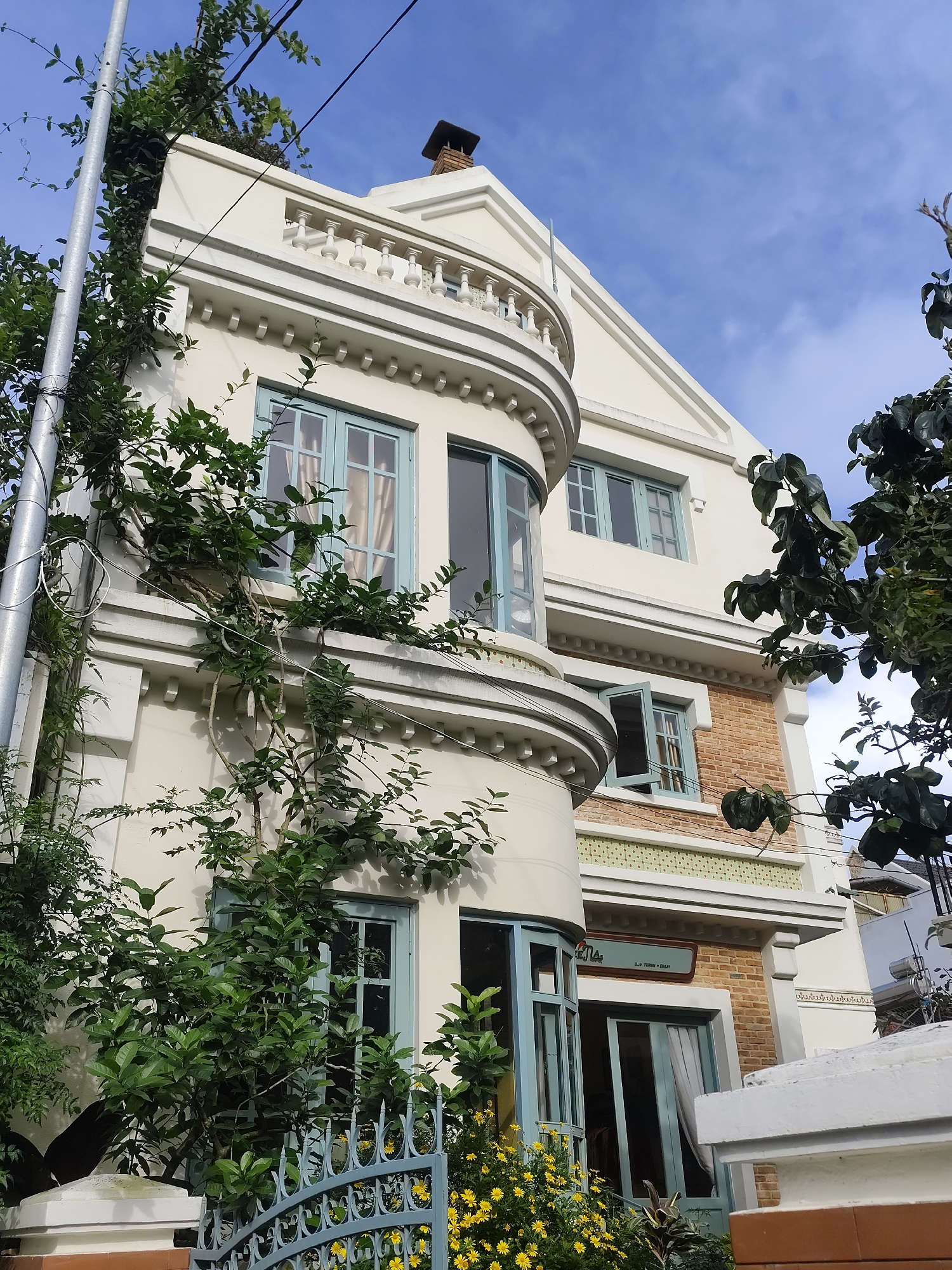 This is an eye-level, medium shot of a cream-colored building in Da Lat, Vietnam. The building has light blue-framed windows and is partially covered in lush green foliage. A tall, grey utility pole with wires stands to the left of the building. In the foreground, a blue wrought-iron fence encloses a garden with yellow flowers and more greenery. The sky above is blue with some wispy clouds. The building appears to have a brick section on its upper floors, and a sign with text, potentially indicating a business or residence, is visible near the ground floor entrance.