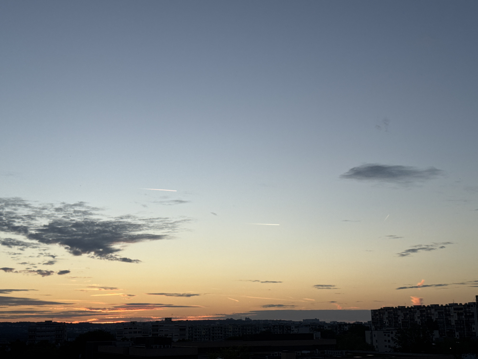 The image depicts a cityscape at dusk, likely in an urban setting such as Unknown, Unknown. The sky transitions from a pale blue at the top to warm hues of yellow and orange near the horizon, indicating the presence of sunset or sunrise. Several thin, white contrails from aircraft are visible across the sky. Scattered clouds, some appearing dark and textured, are present in the upper and middle sections of the image. Below the sky, a silhouette of city buildings is visible. The buildings are varied in height and design, with some appearing to be apartment blocks or office buildings. The foreground is dominated by the cityscape, with the sky taking up the majority of the frame. The overall atmosphere is calm and serene, characteristic of twilight.