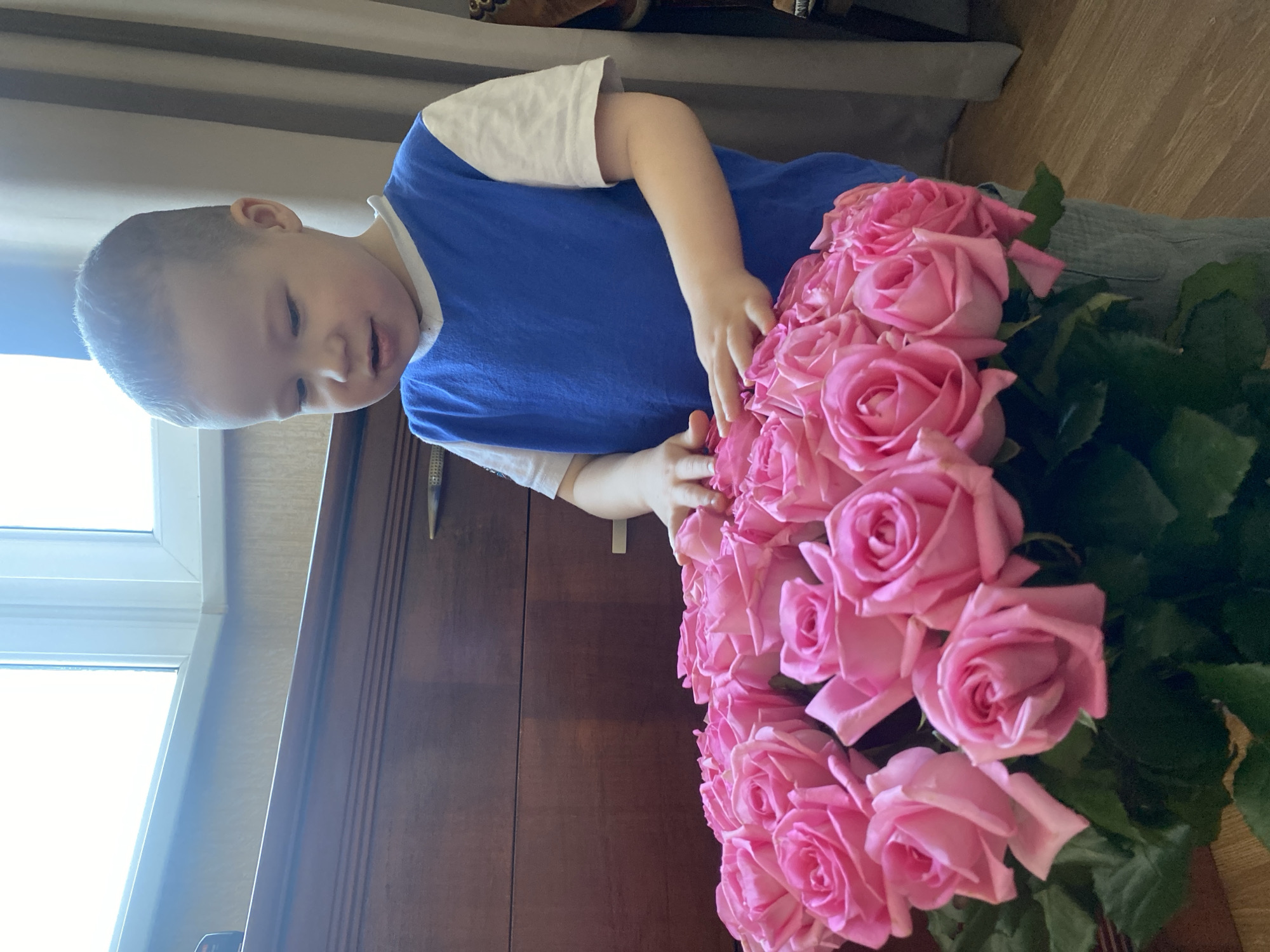 A child with fair skin and short, light brown hair stands next to a large bouquet of pink roses. The child is wearing a blue t-shirt with white sleeves. The bouquet appears to be placed on a dark wooden surface, possibly a table or cabinet. A window is visible to the left. The location is stated as Sochi, Russia.