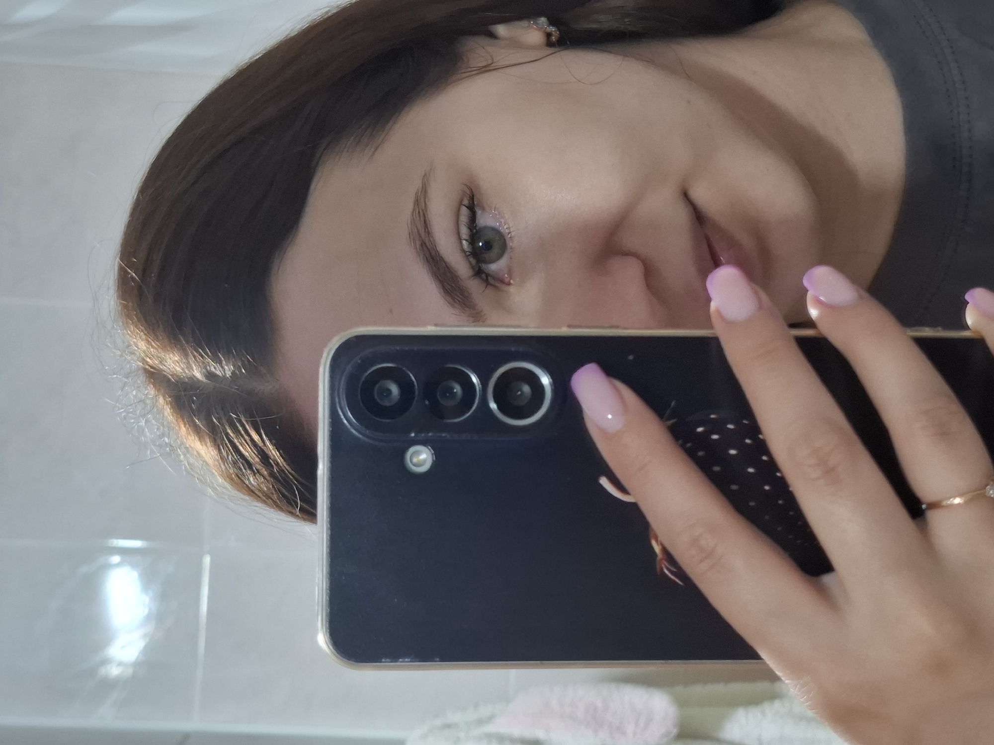 A woman with brown hair is holding a smartphone in her right hand to take a selfie in what appears to be a bathroom. The phone is black with three visible camera lenses on the upper left side of the back. Her nails are painted with a French manicure style using a pale pink base and a purple tip. She is wearing a ring on her ring finger. The background consists of what appears to be a tiled wall, possibly in a bathroom, and a blurred white object below the phone. There's no information in the image to confirm the location in the city of Chaikovsky, Russia.