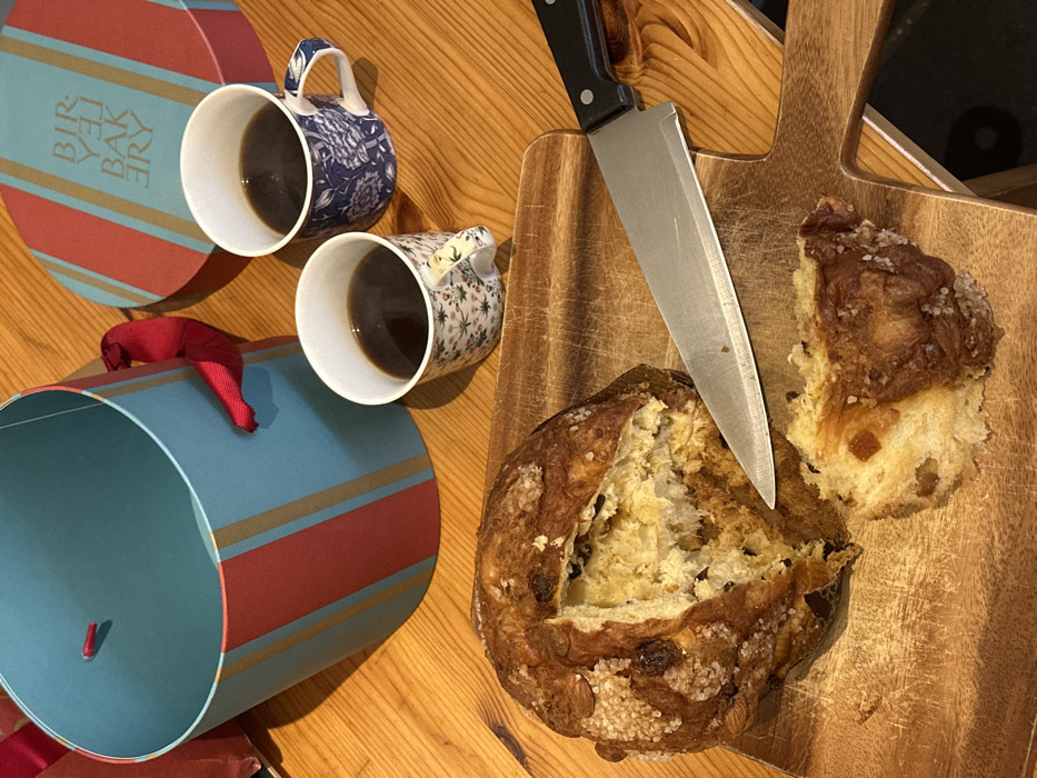 An overhead shot displays a partially consumed panettone and two mugs of coffee arranged on a wooden table. A large, round panettone, featuring a sugared crust with almonds, raisins, and candied fruit, rests on a light-colored wooden cutting board. A section of the panettone has been separated, exposing its soft interior. A chef's knife with a black handle and silver blade lies on the board next to the panettone. Two ceramic mugs containing dark liquid, one with a blue floral pattern and the other with a multi-colored floral design on a white background, are present. In the background, a teal-colored cylindrical box with red and gold stripes is open and empty, alongside its matching lid. The lid displays the mirrored text 'BIRLEY BAKERY'. The scene depicts a meal in London, United Kingdom, likely related to Birley Bakery, which is located in Mayfair.