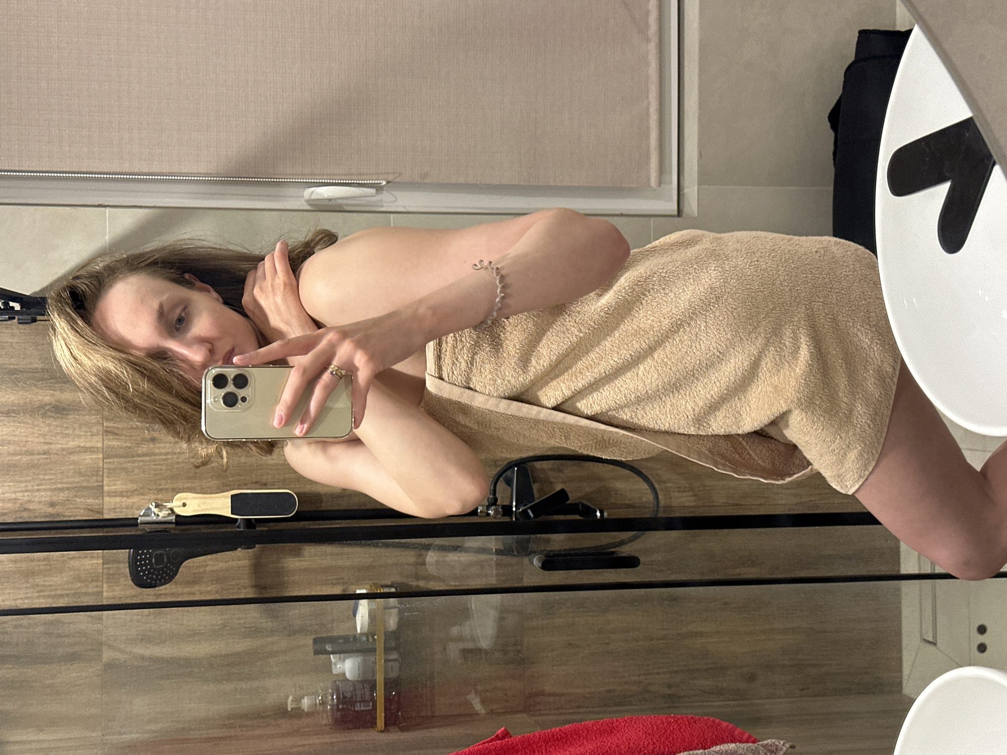 The image shows a woman in a bathroom taking a selfie in a mirror. The woman is blonde and appears to be wearing a towel. She is holding a gold iPhone. In the background there is a window with a shade, a sink, and a shower with a foot file hanging in it. There are also several bottles of products on a shelf. The bathroom is of modern design with light beige tiles. I am unable to identify the location as the city of Sepych, Russia.