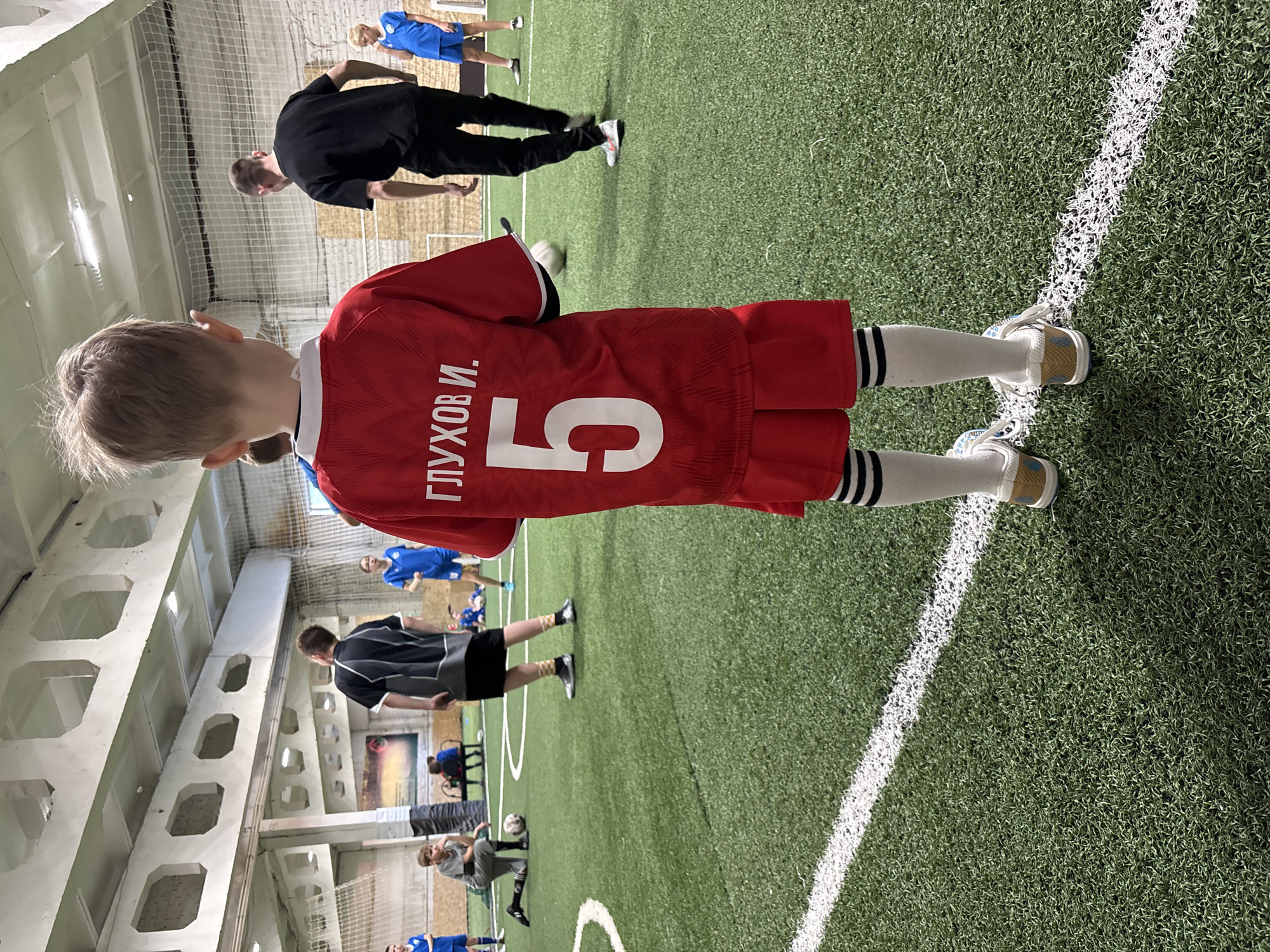 A child in a red soccer jersey with the number "5" and the Russian name "Глухов И." (Glukhov I.) stands on a green artificial turf field marked with white lines. He is wearing white socks with black stripes and white shoes. Other individuals, including adults and children in various sportswear, are scattered across the field, engaged in soccer-related activities. The scene appears to be an indoor soccer facility, possibly in Chaikovsky, Russia, based on the name on the jersey and the general indoor athletic setting. The ceiling features a grid-like pattern of supports and lights. A net is visible along the side of the field.