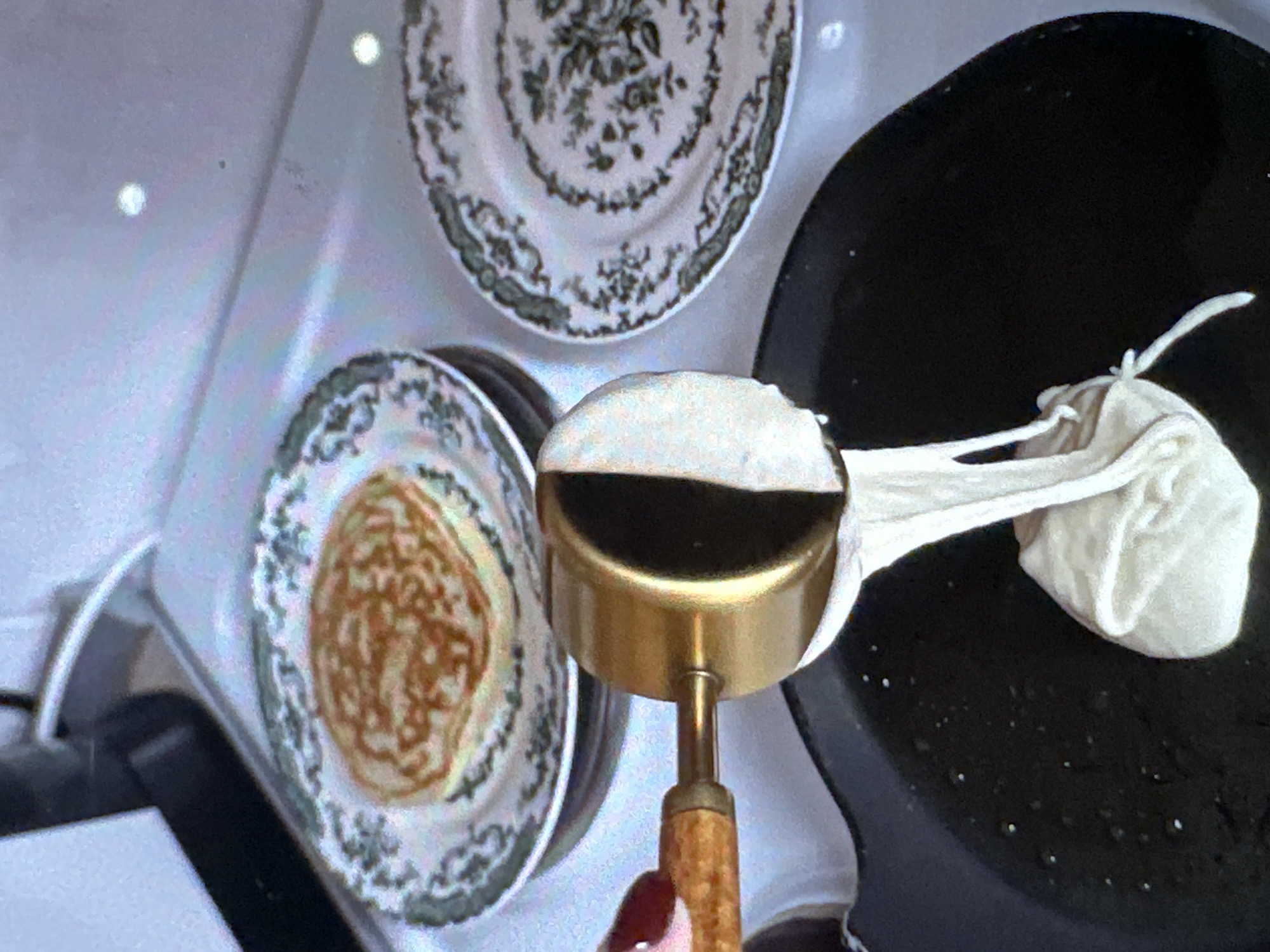 The image depicts a cooking scene. A brass-colored utensil is positioned over a black cooking pan, possibly a skillet or griddle. A white fabric, appearing like a cloth bag or pouch, is attached to the utensil and hangs over the edge of the pan. A white substance, possibly batter or dough, is being squeezed or dispensed from the fabric pouch into the pan. In the background, there are two stacked plates with floral patterns, one holding a cooked pancake or crepe.
There is no data to confirm the location of this image being in the city of Yerevan, Armenia.