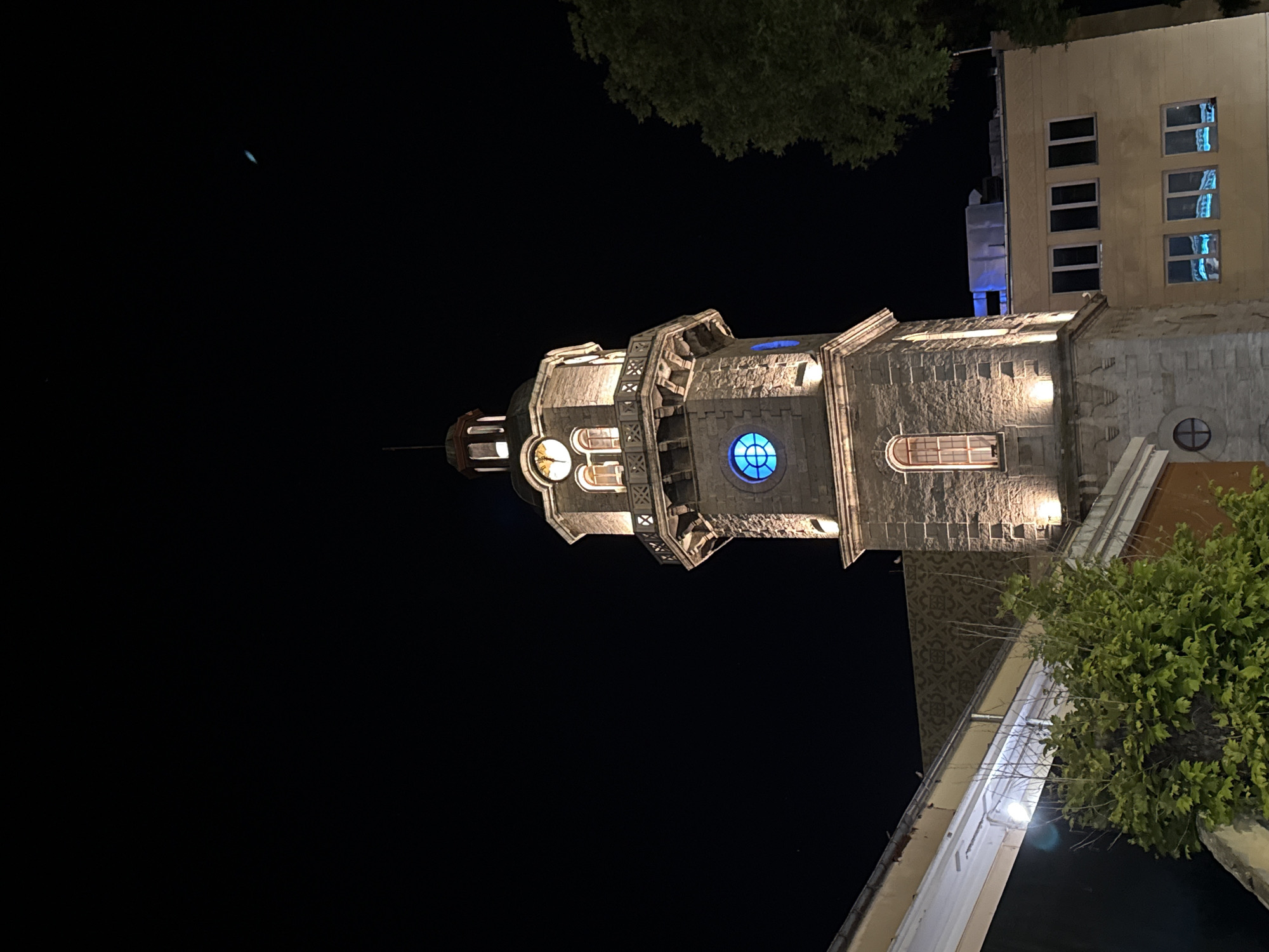 The image shows a stone clock tower at night, illuminated by warm lights. The tower has a clock face with Roman numerals and a small spire on top. Below the clock, there is a balcony with decorative railings. A round window with blue light is visible on the tower. To the left of the tower, a building with a yellow facade and dark windows is partially visible, along with some greenery. To the right, the branches of a large tree obscure the view. In the background, a modern building with multiple windows is seen, with a blue light reflecting in some of the windows. The sky is dark and clear, with a few faint distant lights. This is the clock tower of the Saints Constantine and Helena Orthodox Church in Varna, Bulgaria.