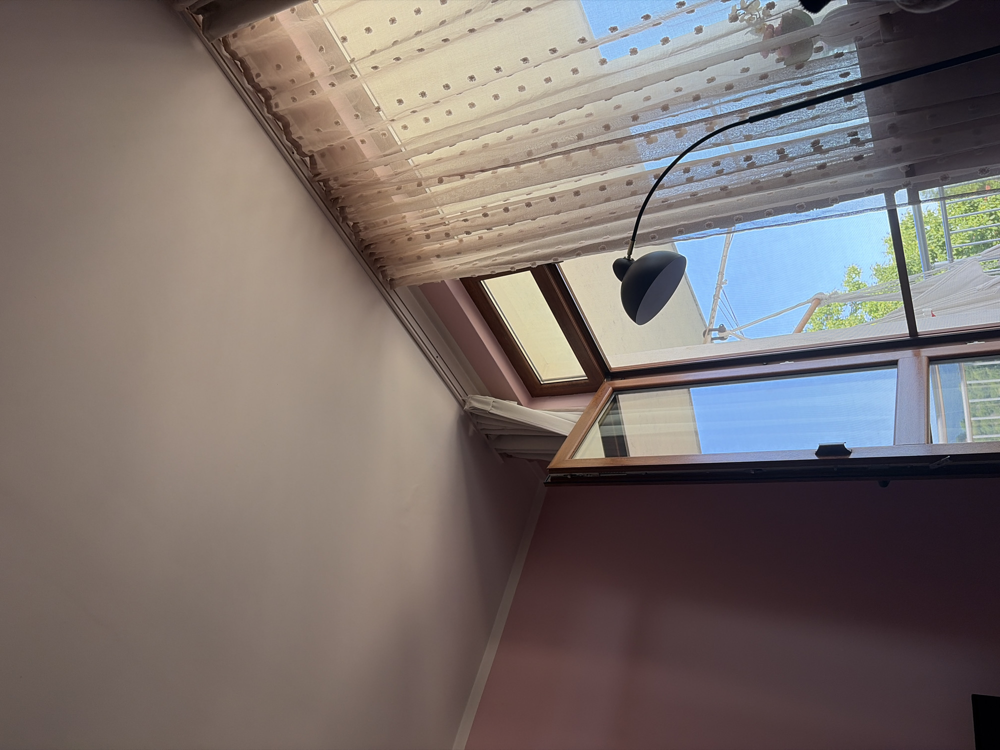 This is a medium shot, indoor, eye-level, vertical, slightly low-angle photograph of a room interior in Tsarevo, Bulgaria. The room has a pink wall on the left and a large open window with curtains on the right. A black arc lamp is positioned in front of the window. The ceiling is white and shows a slight shadow. The window has a wooden frame and is partly open, revealing a bright blue sky and some trees outside. The curtains are sheer, light-colored, and decorated with small, dark floral patterns. They are drawn open to the right. The lamp has a curved neck and a black, dome-shaped shade. The overall impression is a bright, airy room.