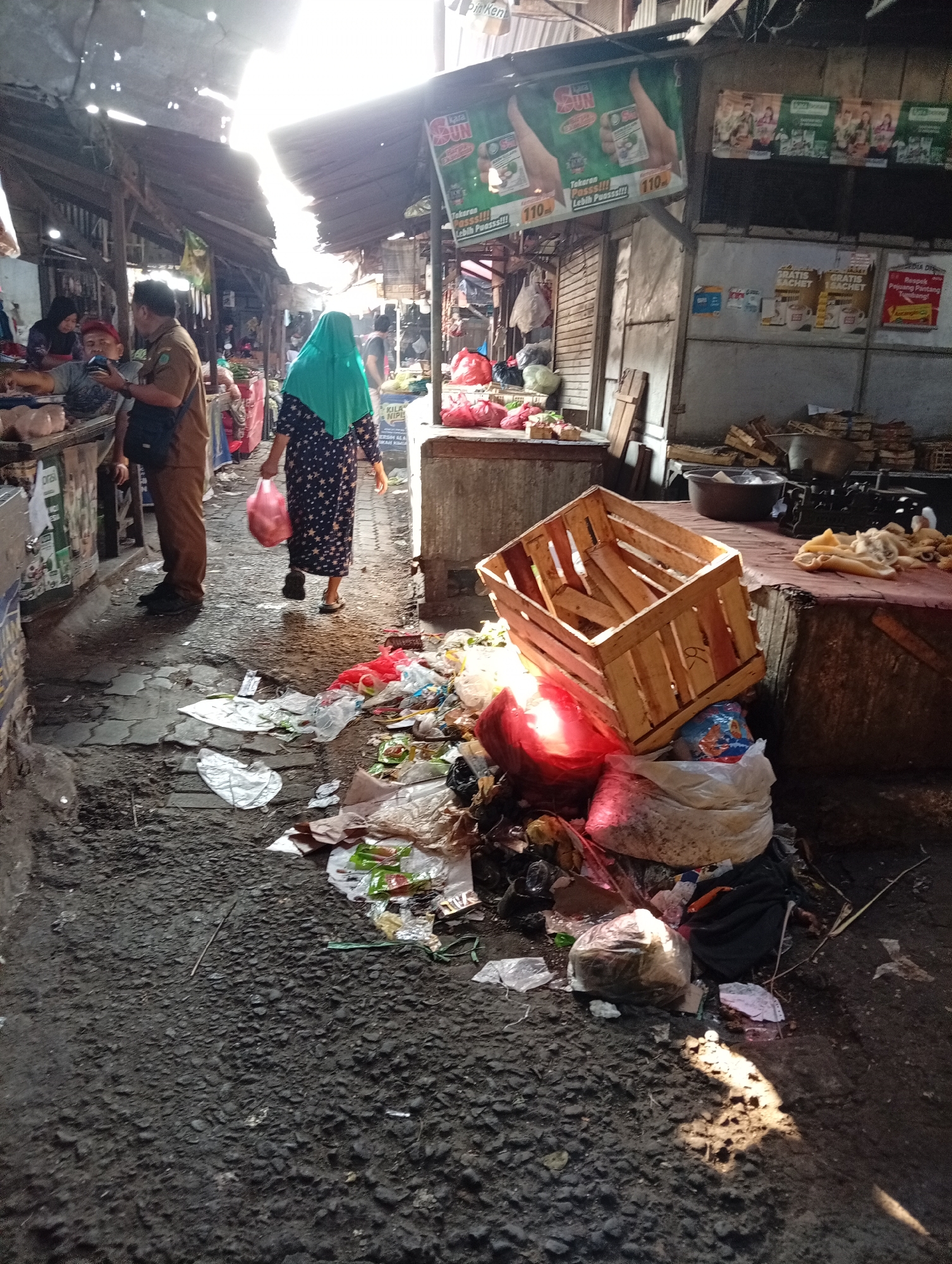 The image depicts a bustling marketplace in Larangan, Indonesia, characterized by a stark contrast between commercial activity and accumulated waste. The narrow alleyway is lined with stalls, some open and others closed. Sunlight streams through gaps in the overhead tarpaulin roofing, illuminating dust motes and casting long shadows.
In the foreground, the ground is heavily littered with a variety of refuse. This includes plastic bags, crumpled paper, food wrappers, and what appear to be discarded food items. A large, overturned wooden crate sits prominently amidst the trash, some of which is red and appears to be the contents of a plastic bag. A bright red spherical object, possibly a balloon or a similarly shaped item, is also visible within the debris.
Several individuals are present, engaged in market activities. On the left, a man in a brown uniform, possibly a market official, is interacting with another person. A woman wearing a teal hijab and a dark dress patterned with white stars walks away from the viewer, carrying a pink plastic bag. Other figures are visible further down the alley, their activities indistinct.
The stalls themselves are varied. Some display goods, while others are stacked with wooden crates or covered with tarpaulins. Advertisements are plastered on some of the stall fronts, including ones for "SUN" and what appear to be food or beverage products. The overall impression is one of a vibrant, yet somewhat disorganized and unclean, urban market environment.