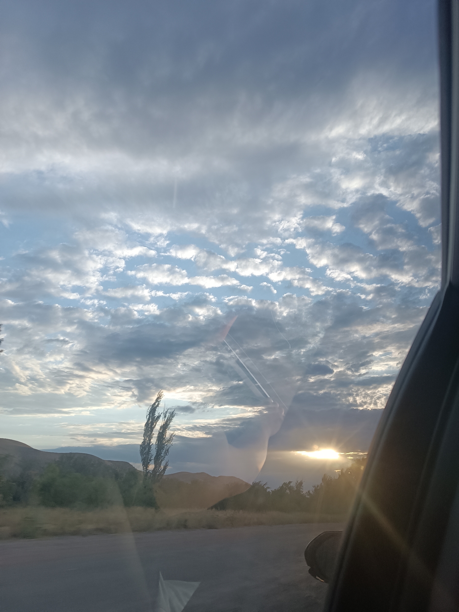 The image depicts a view from a vehicle looking out at a landscape under a cloudy sky. The scene captures a stretch of asphalt road in the foreground, bordered by dry, yellowish vegetation and low, rolling hills in the distance. Two tall, slender trees with sparse foliage stand prominently in the mid-ground, silhouetted against the horizon. The sky is filled with a variety of clouds, ranging from dark, heavy formations to lighter, scattered patches that allow sunlight to break through. The sun is visible as a bright, glinting orb near the right side of the frame, casting a strong glare and lens flare across the image. The overall impression is of a rural or semi-rural environment, possibly during late afternoon or early morning given the quality of the light. The location is identified as Sasyk-Bulak, Kyrgyzstan. There are no people or specific actions visible in the image, and the focus is on the natural landscape and atmospheric conditions.