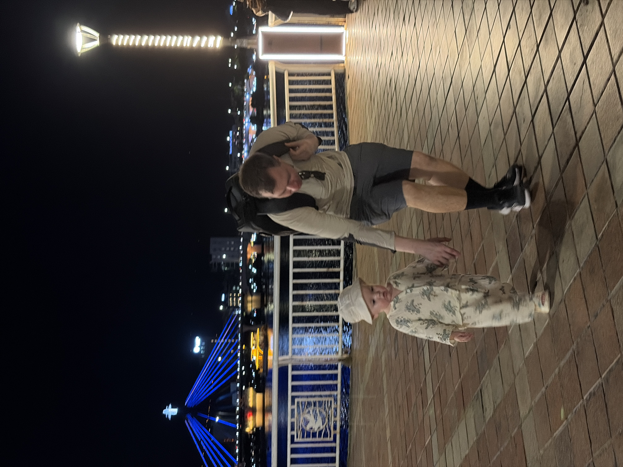 This image depicts a nighttime scene on a well-lit urban promenade, likely in Đà Nẵng, Vietnam. In the foreground, a man and a young child are captured on a patterned paved walkway.
The man, positioned towards the center-right, wears a light-colored long-sleeved shirt, dark shorts, dark socks, and athletic shoes. He carries a dark backpack and has sunglasses tucked into his collar. He is looking down and reaching his right hand towards the child. The child, a toddler or small child, stands to the man's left, dressed in a light-colored, patterned long-sleeved outfit and a bucket hat. The child looks towards the viewer.
The walkway features a distinctive diamond-shaped tile pattern. To the left of the subjects, a white railing with blue accents and decorative panels separates the promenade from what appears to be a body of water, reflecting distant lights.
The background is dark, indicating night. Various artificial lights illuminate the scene. A tall, decorative light pole with a spiral of bright white lights is prominent on the far left. Further in the distance, the iconic Dragon Bridge, illuminated with striking blue lights, spans across the background, a strong location cue for Đà Nẵng. Other city lights from buildings are also visible along the horizon. The overall impression is a pleasant evening stroll or outing. There is no visible text in the image.