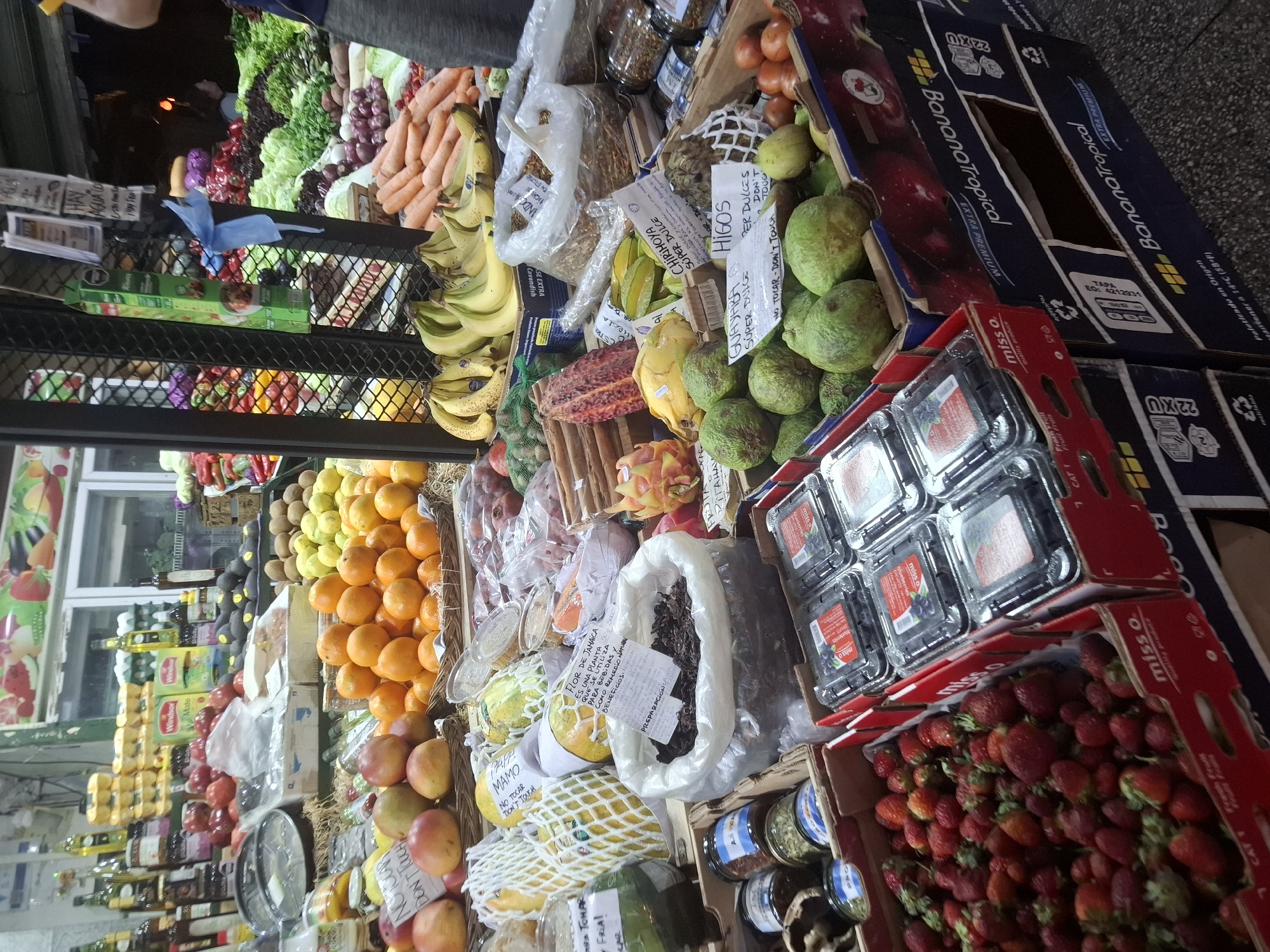 This is a photograph of a produce stand in Buenos Aires, Argentina. The stand is overflowing with a colorful array of fruits and vegetables including carrots, grapes, lettuce, bananas, oranges, mangos, kiwis, strawberries, blueberries, guavas, figs, and cherimoyas. Signage in Spanish indicates the names of the fruit and prices. There are jars of preserved goods, bottles of oil, and other items stacked on the shelves behind the produce. One person is partially visible at the top of the frame. The products are displayed on multiple levels. Some of the boxes are labeled "BananaTropical".