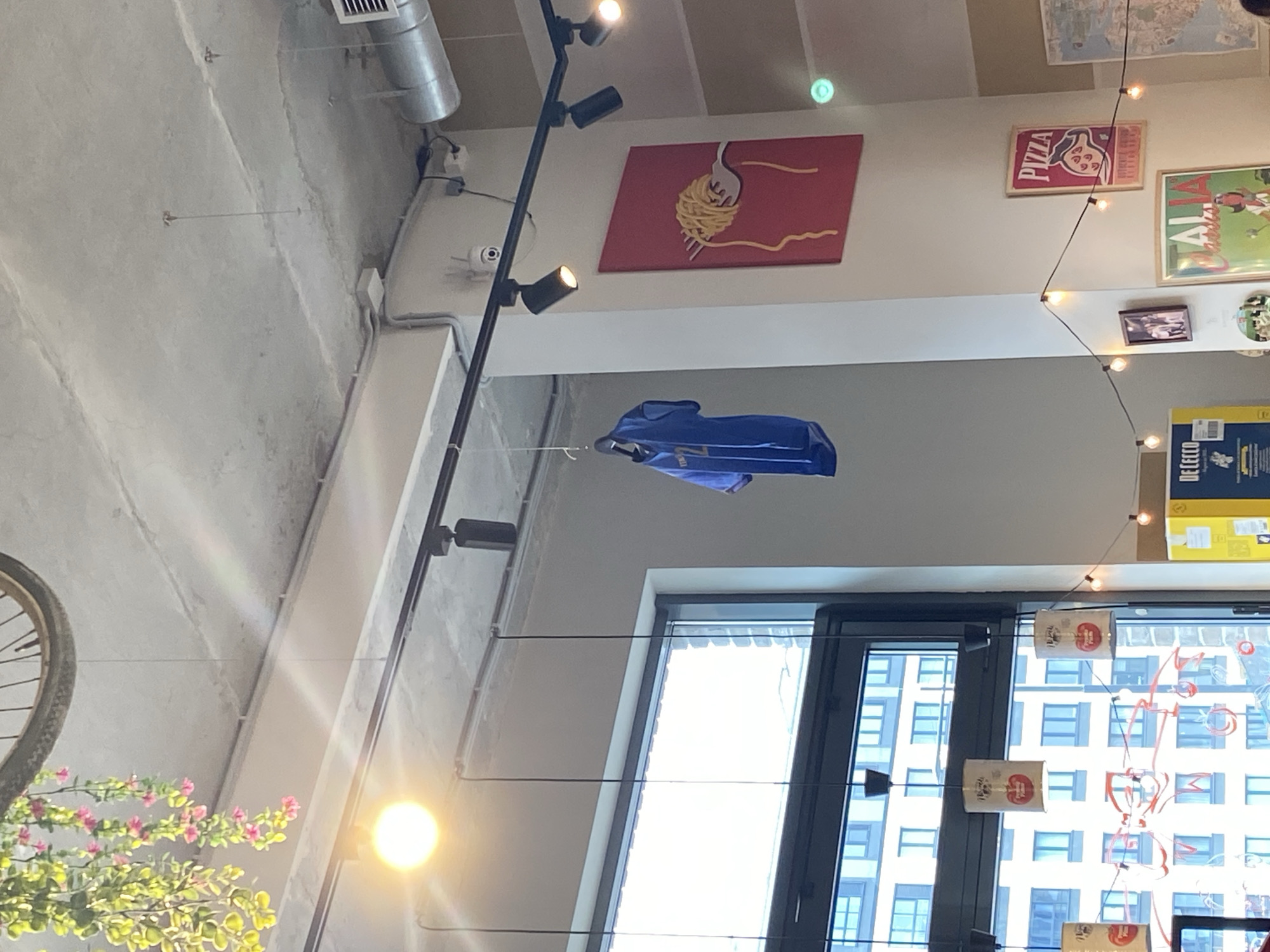 The image depicts the interior of a building with a high ceiling and various decorations. A blue jacket is suspended in mid-air by a thin wire. Decorative elements include a red panel with a spaghetti and fork graphic, several posters, and string lights with small bulbs. A bicycle wheel and plants are visible in the lower-left corner. A window reveals an exterior cityscape with high-rise buildings. The location is not identifiable as Izhevsk, Russia from visual details alone.