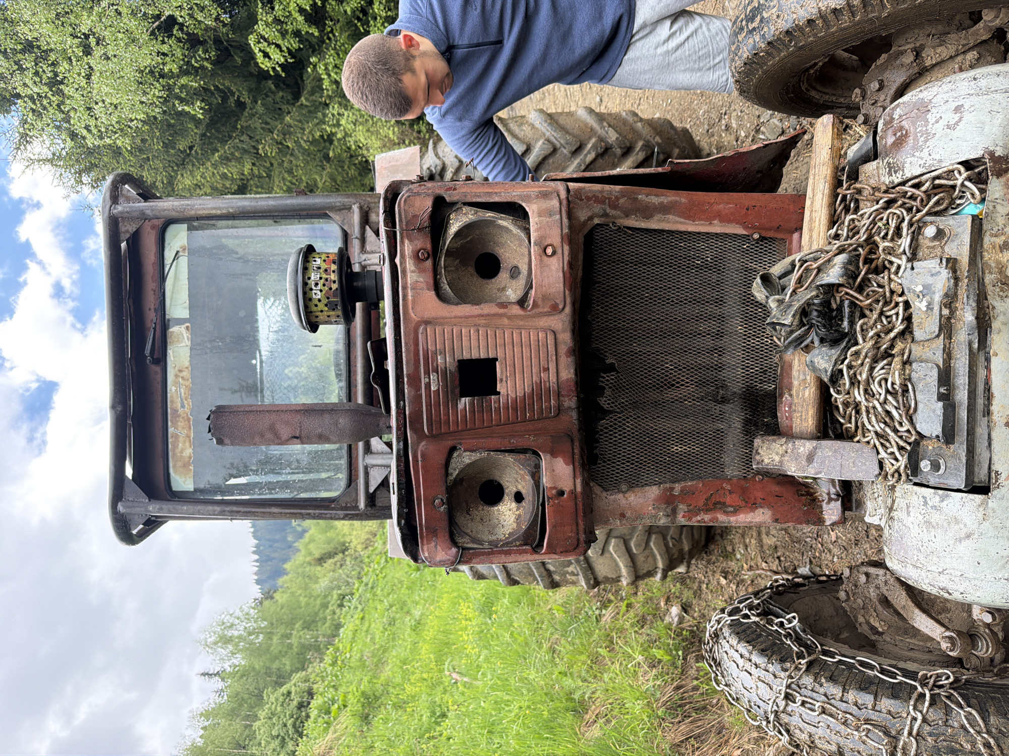 The image depicts a man leaning over the front of a weathered, red tractor. The tractor's headlights are missing, and the grill is visible. Chains are draped over the front of the tractor, near the wheels. The tractor is parked in a rural area with green vegetation and trees in the background. The setting appears to be in a mountainous region, possibly near a forest. Given the location hint, the scene is potentially located in the city of Рилски манастир, Bulgaria. The man is wearing a blue shirt and grey pants, and appears to be inspecting or working on the tractor.